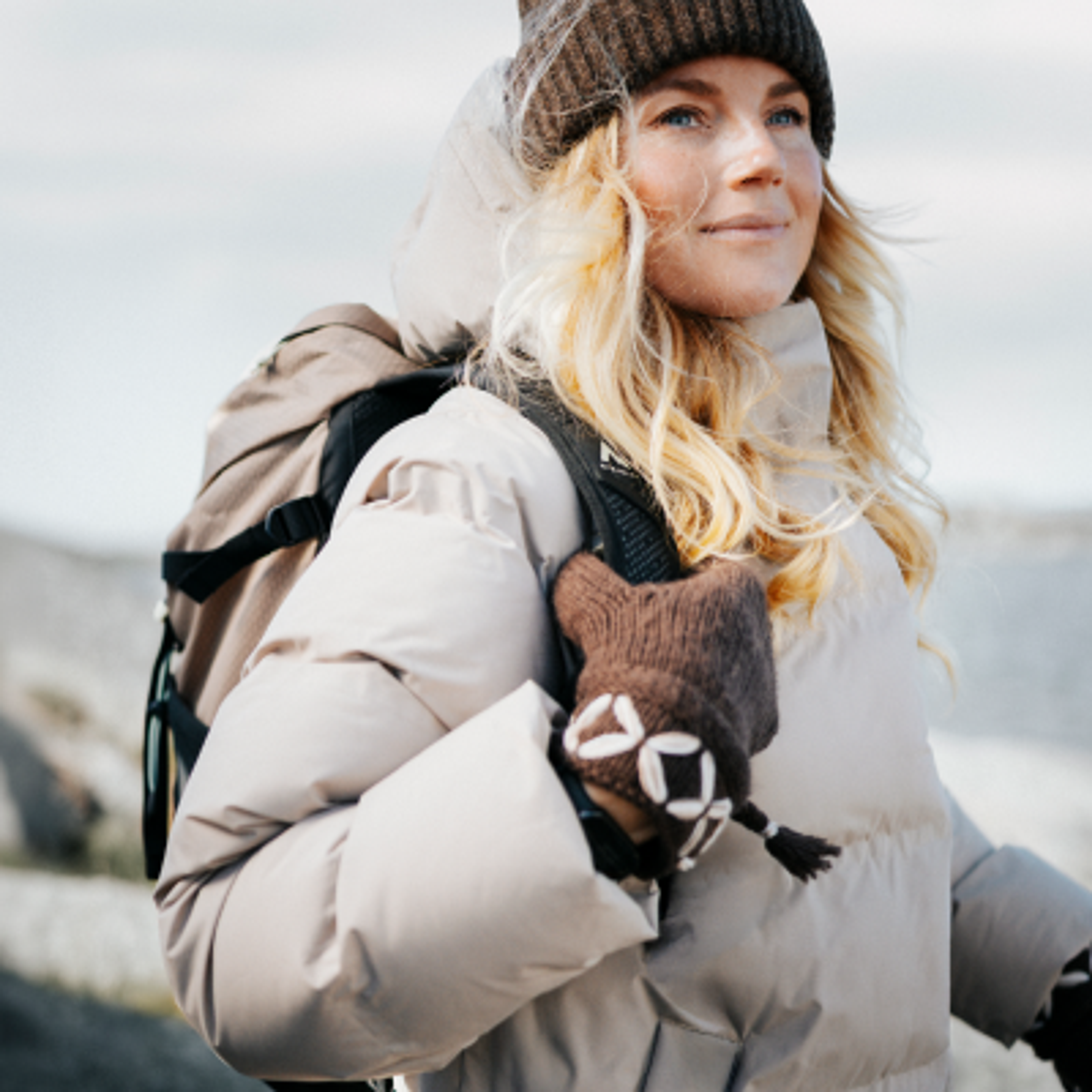 Blonde woman in a brown beanie, beige puffer jacket, and patterned mitten, with a backpack, looking up outdoors.