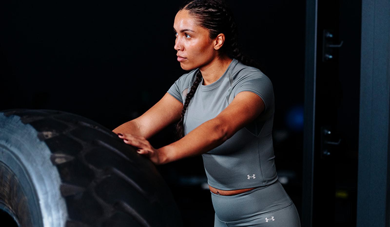 A woman in grey athletic wear pushes a large black tire in a gym.