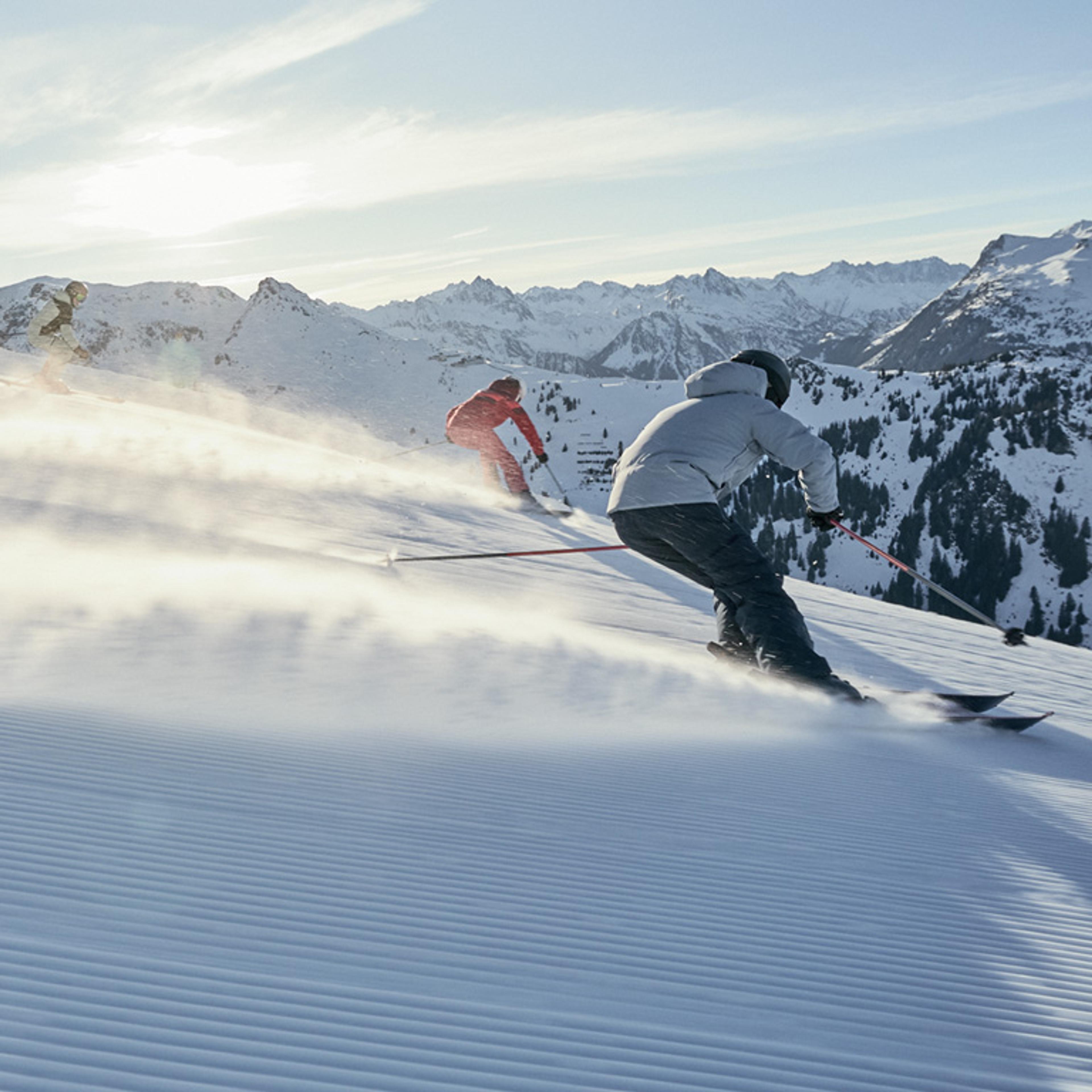 Three skiers descend a sunny, groomed mountain slope, kicking up snow with snow-capped peaks in the background.