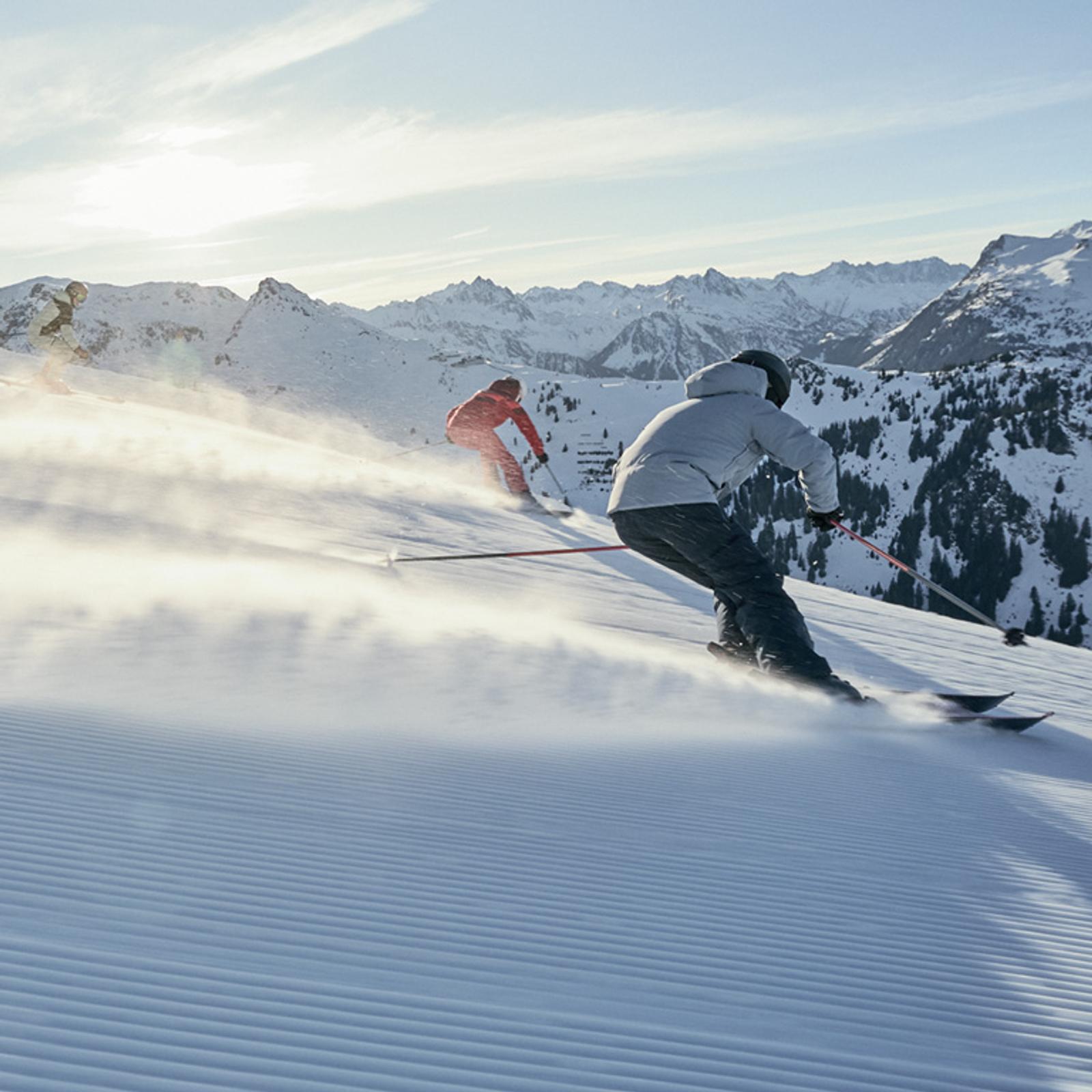 Three skiers descend a sunny, groomed mountain slope, kicking up snow with snow-capped peaks in the background.