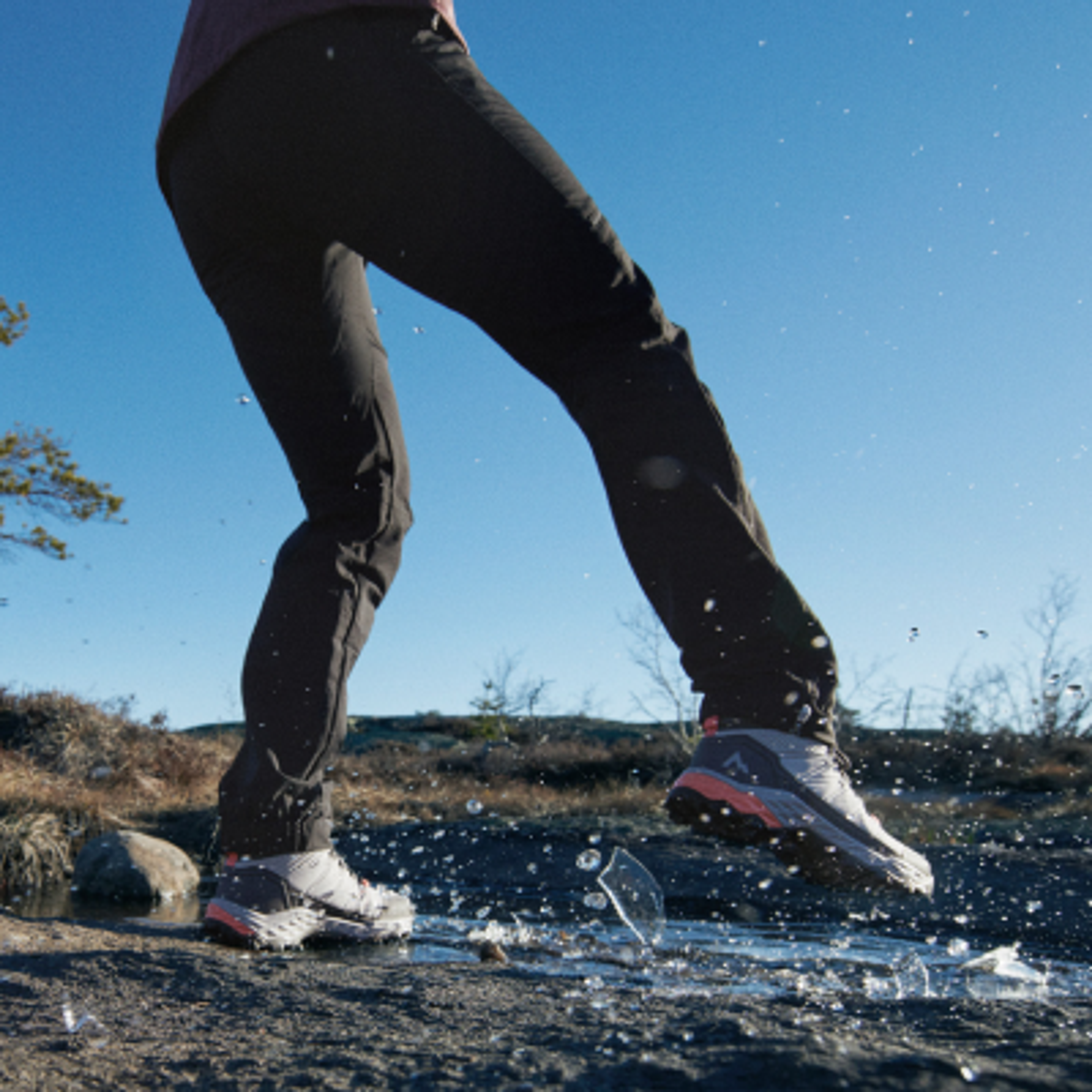 A person's shoe breaks thin ice, splashing water on rocky ground.