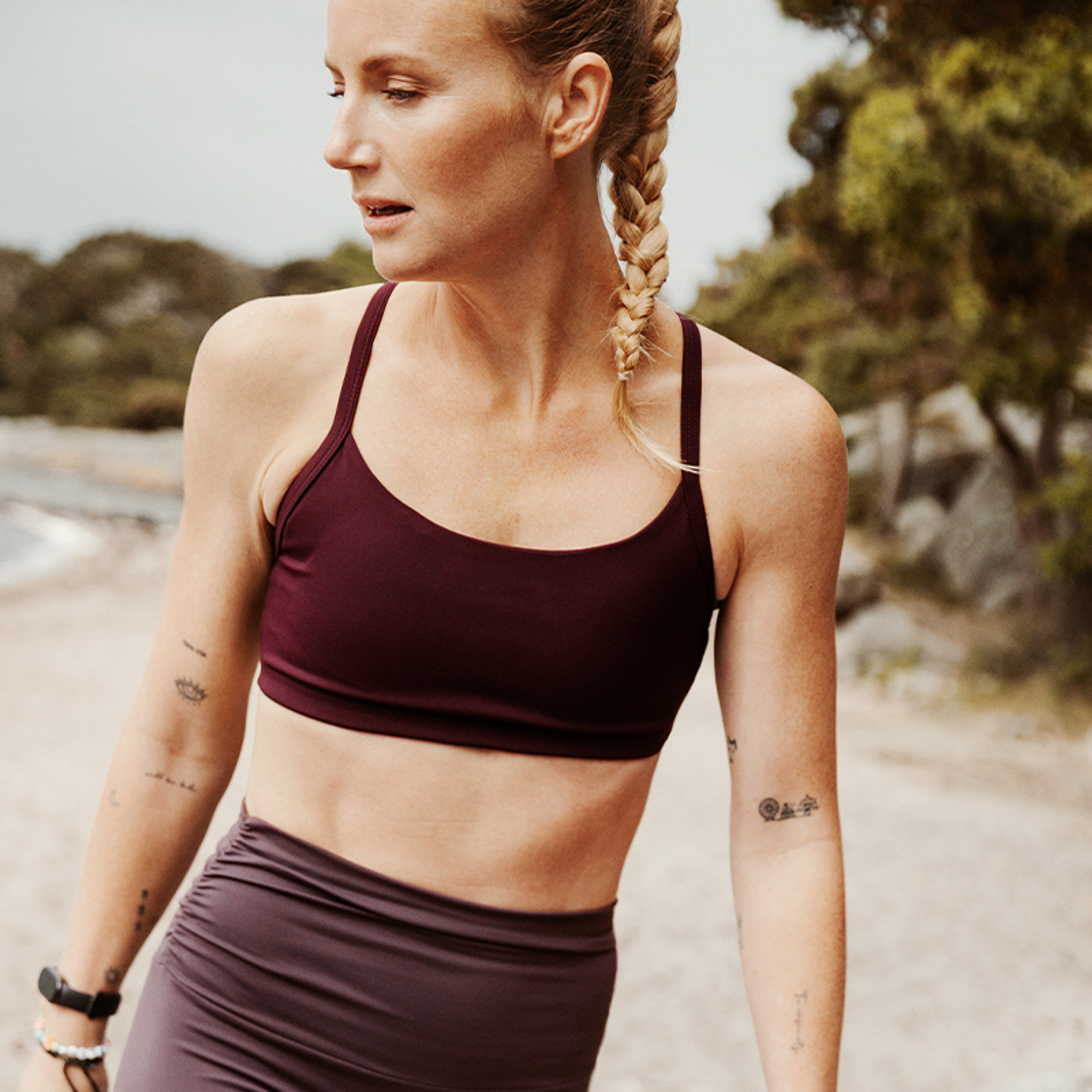 A woman with a blonde braid wears a maroon sports bra and purple leggings, looking left outdoors.
