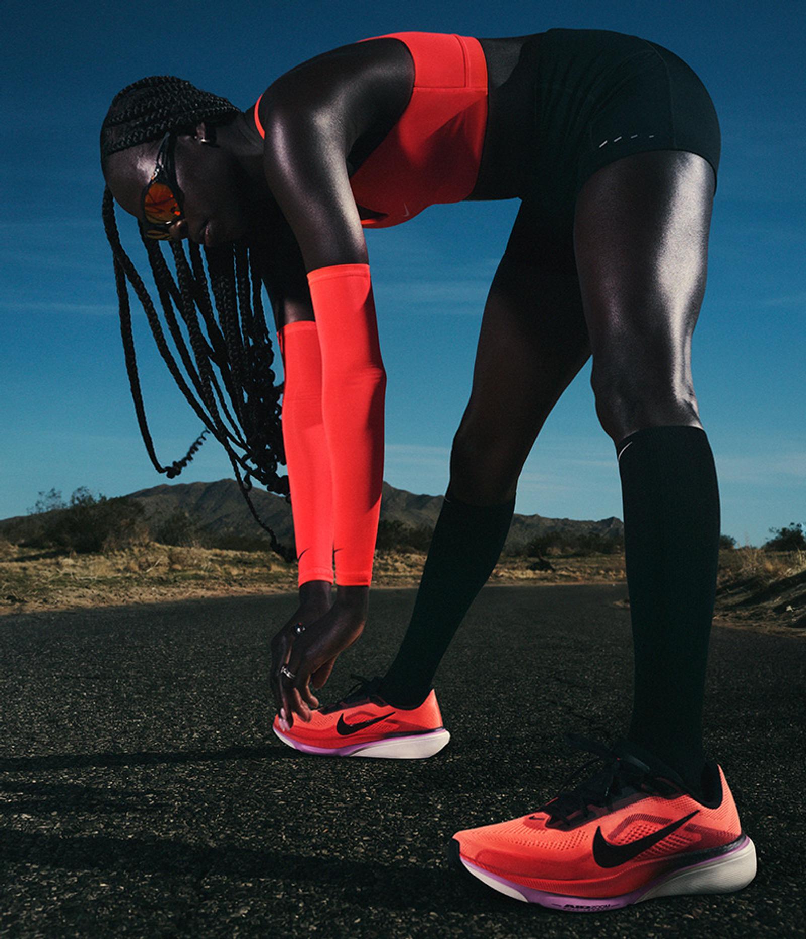 A dark-skinned runner in a red top, black shorts, and red arm sleeves bends to stretch on a desert road, wearing vibrant orange Nike running shoes.