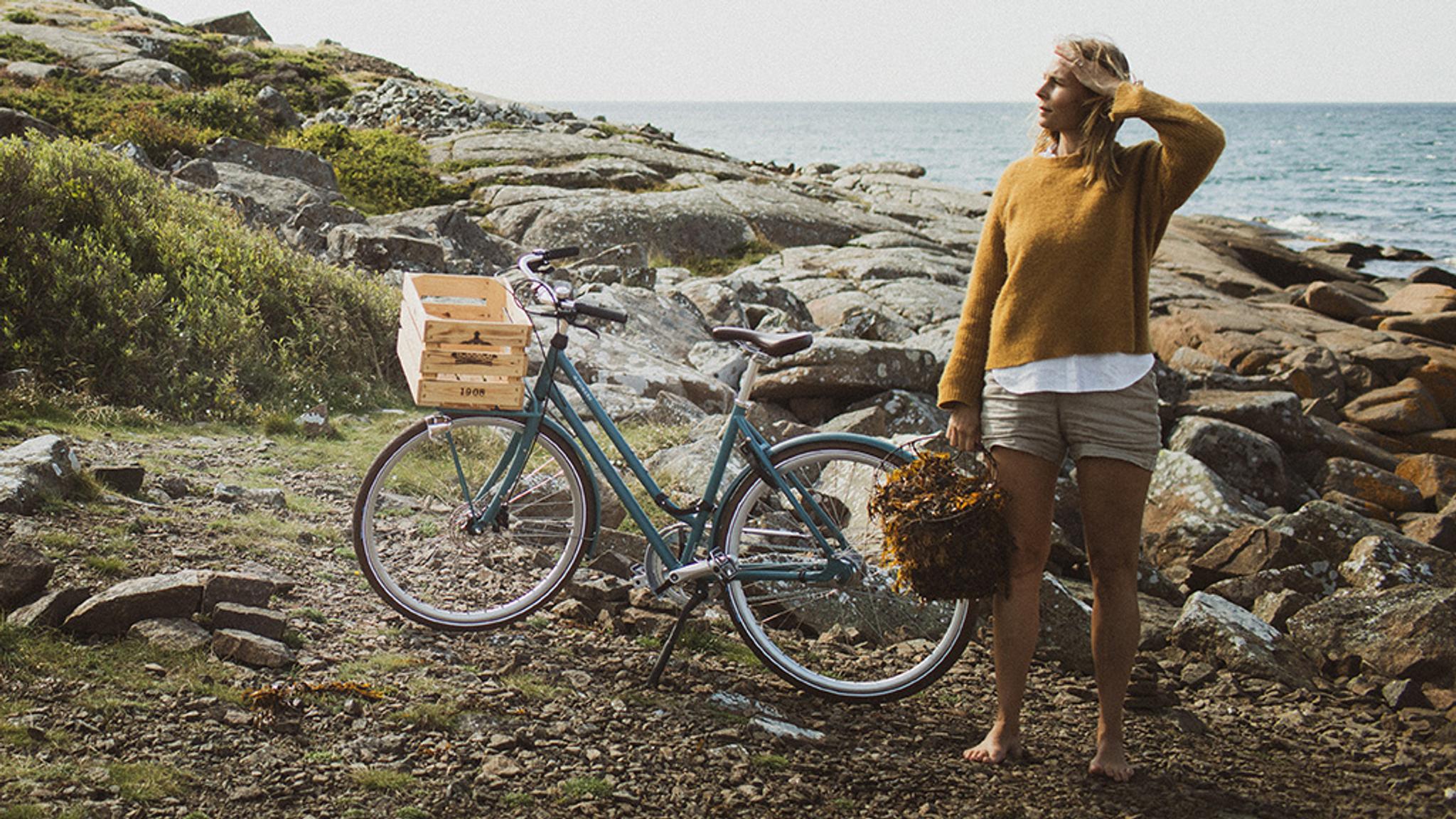 A woman holding a basket of seaweed stands barefoot on a rocky coast next to a bicycle with a wooden crate, looking towards the sea.