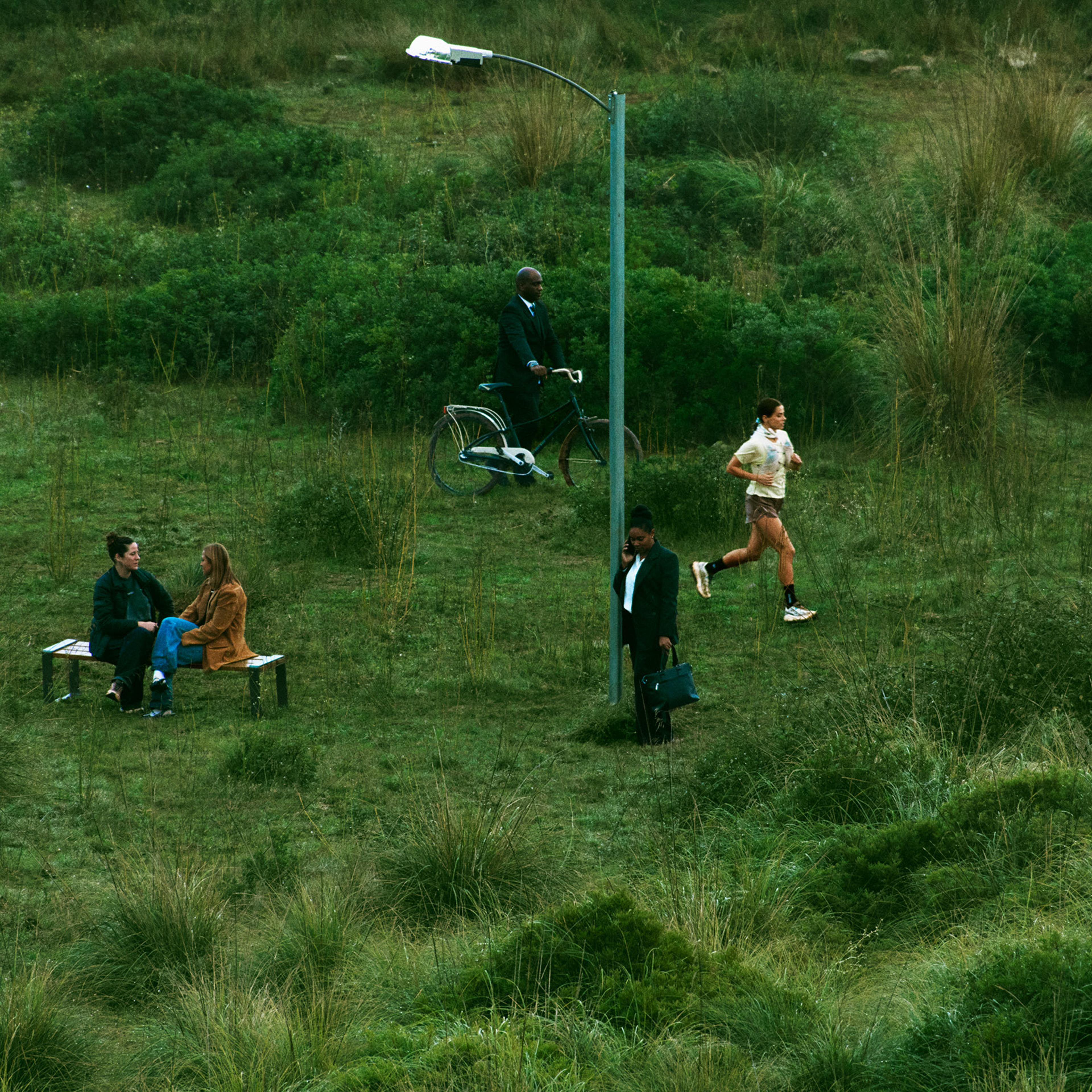 People sit on a bench, cycle, jog, and talk on a phone in an overgrown green field with a lamppost.
