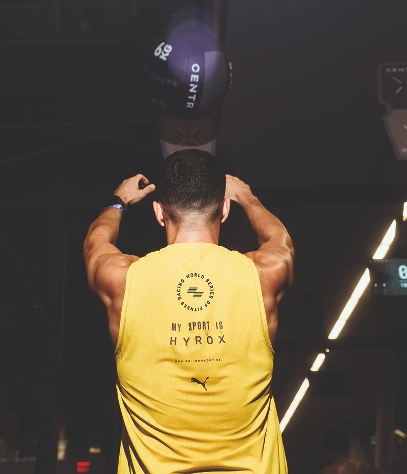 Muscular person in yellow "MY SPORT IS HYROX" tank top looking up at a 6kg wall ball in a gym.