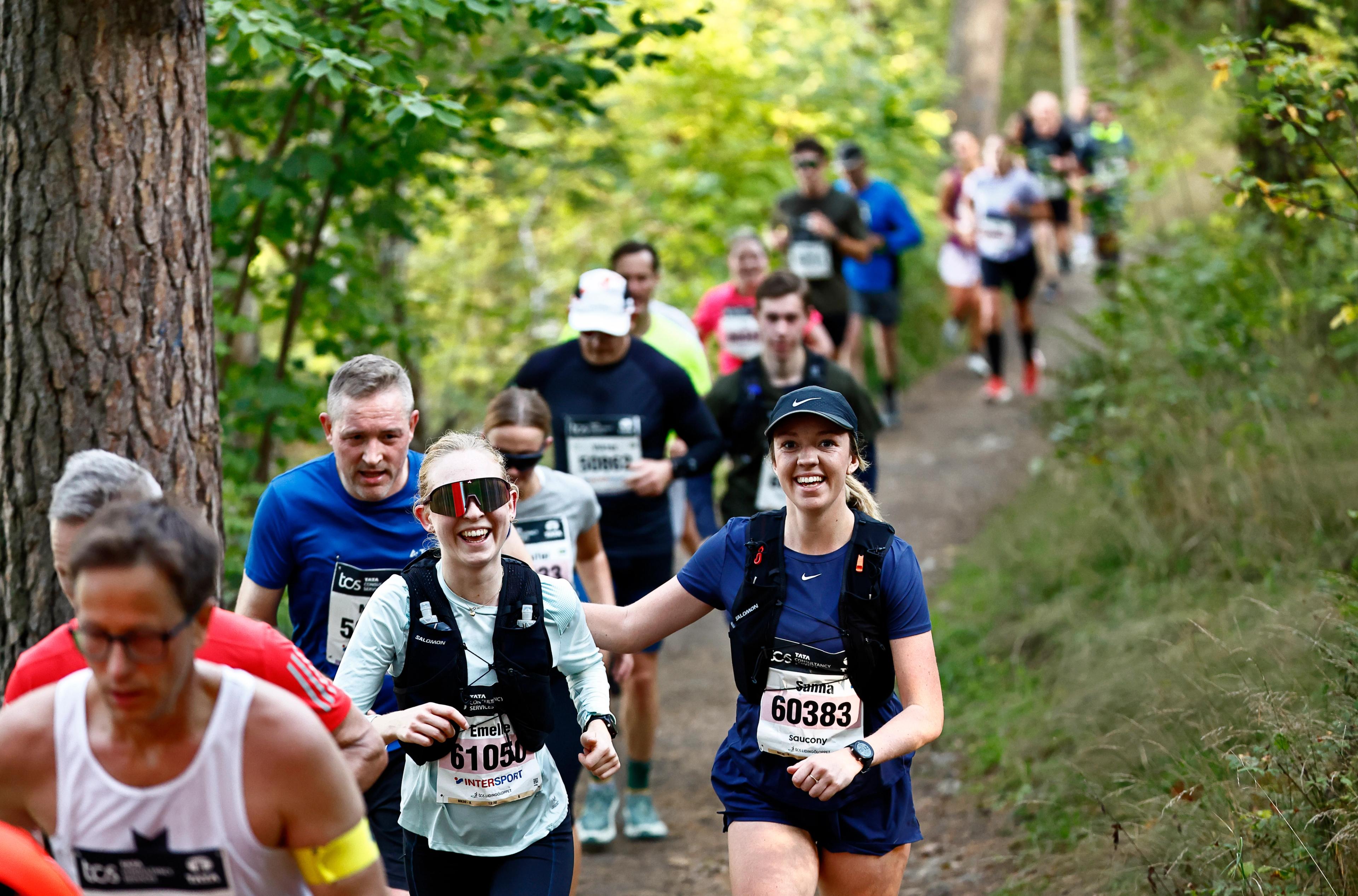 A group of smiling runners ascending a trail through a forest during a race.