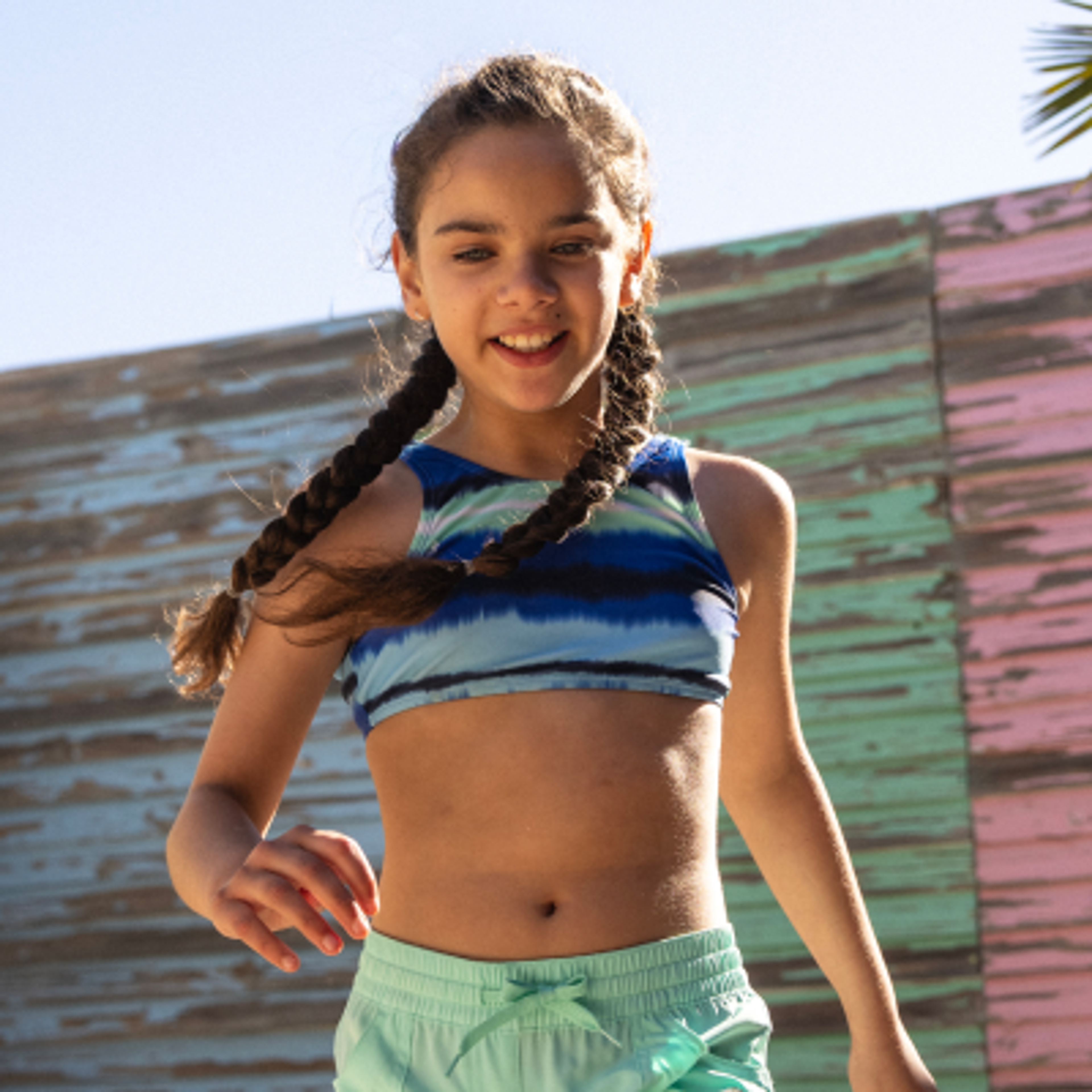 Smiling girl with braids in a blue tie-dye top and mint shorts against a colorful wooden wall.