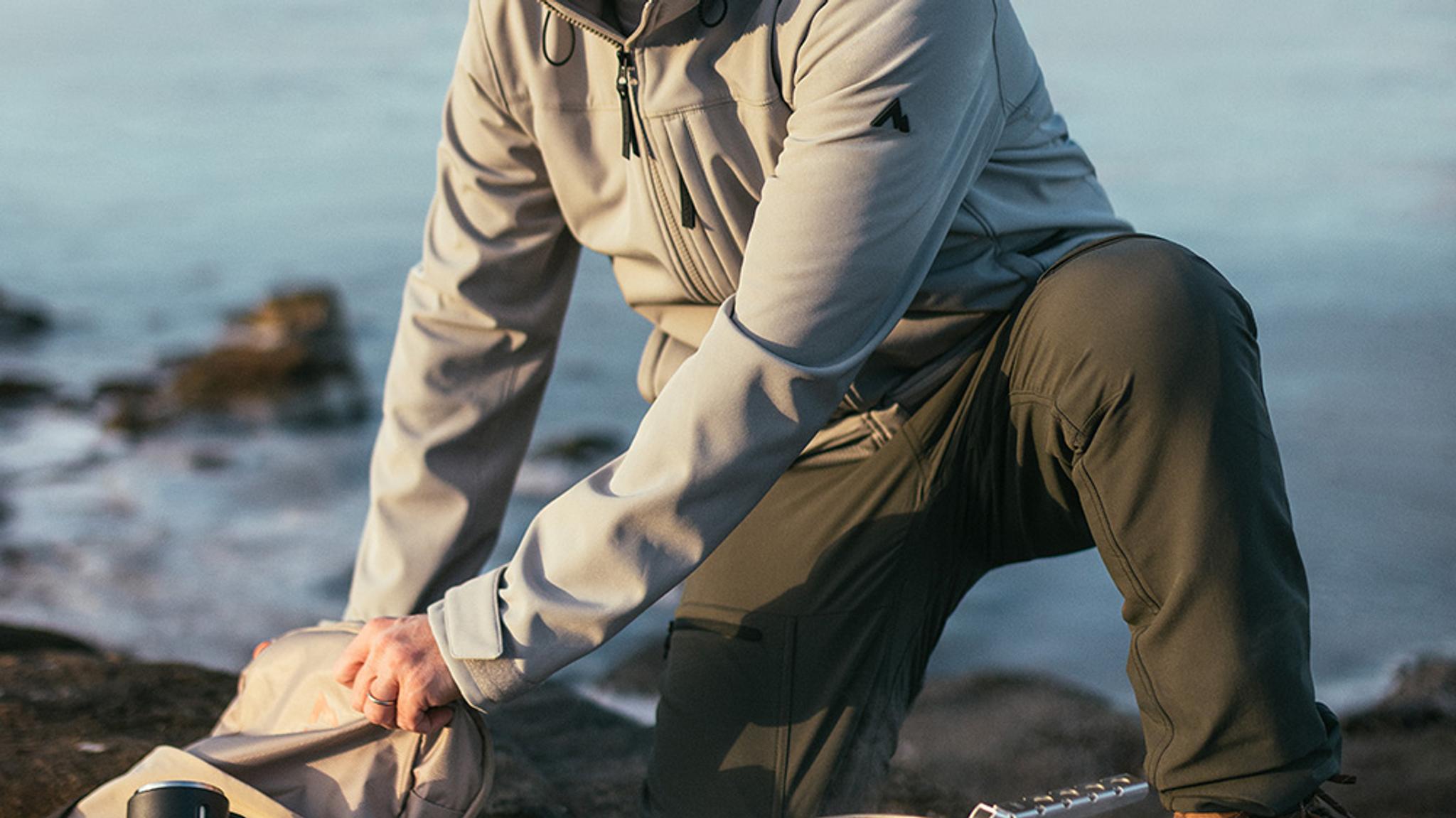 A person wearing a light jacket and dark pants kneels by the water, reaching into a bag.