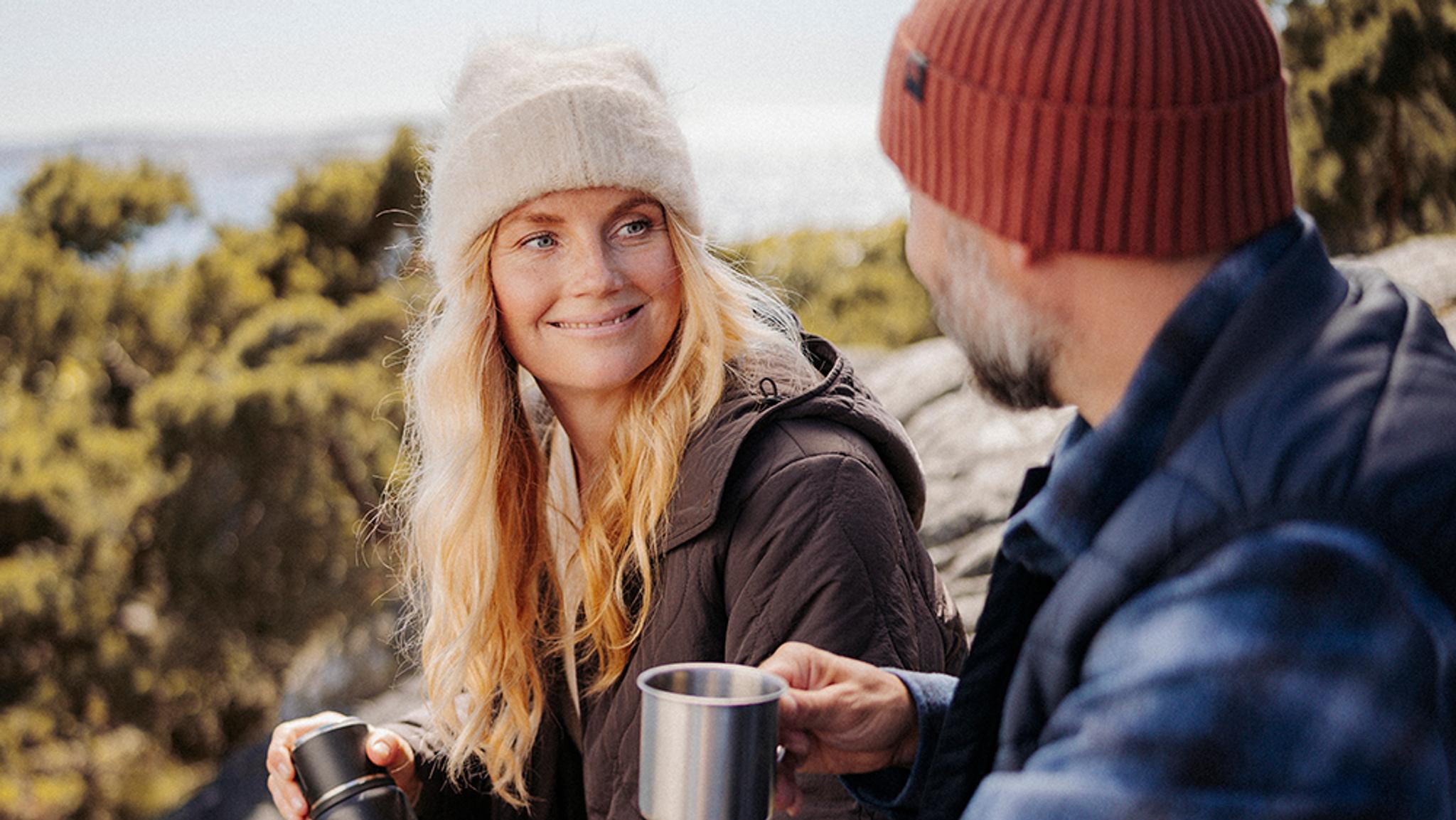 A smiling woman in a beanie looks at a man holding a mug outdoors.