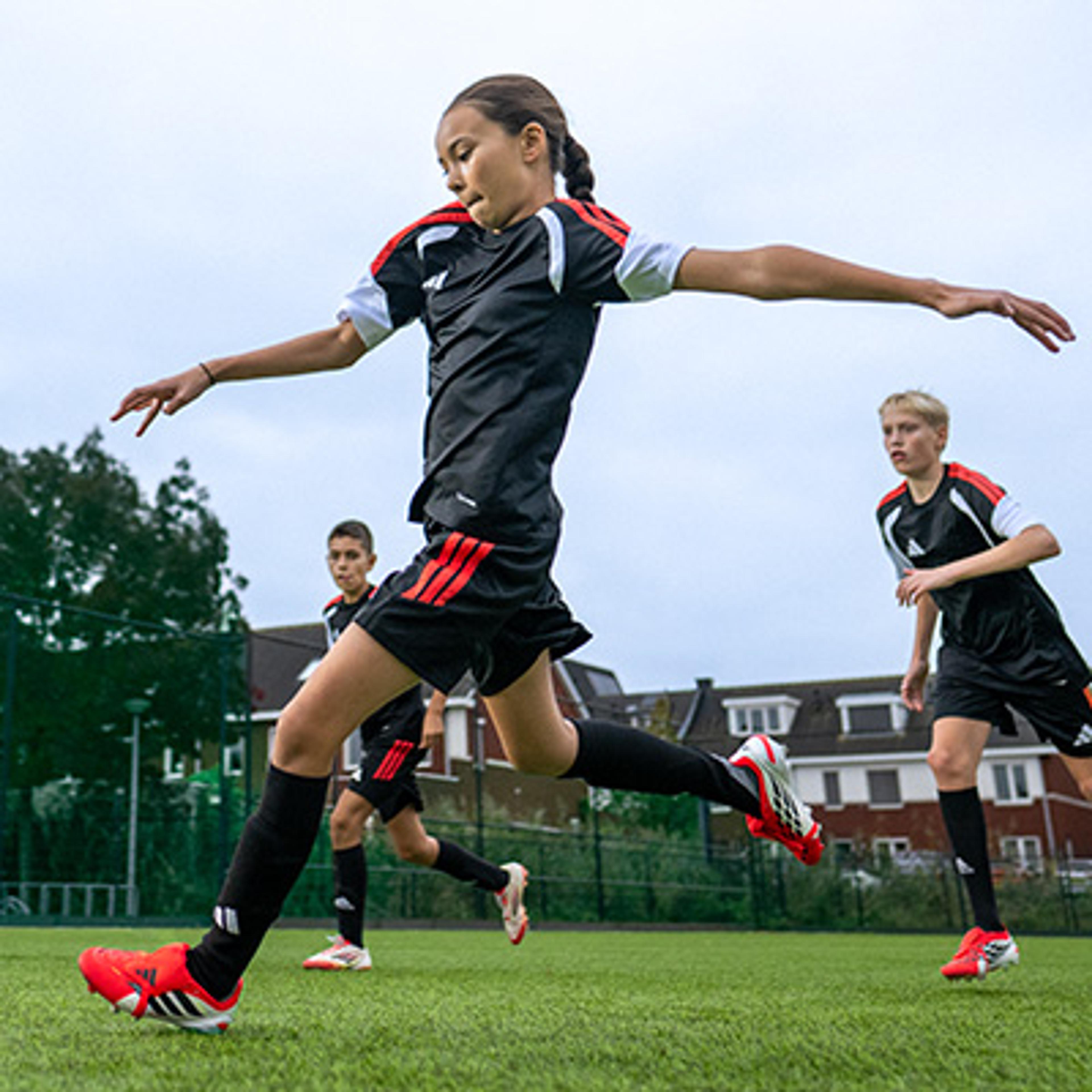 A young female soccer player in a black and red uniform runs on a green artificial turf field, with two other players in the background.