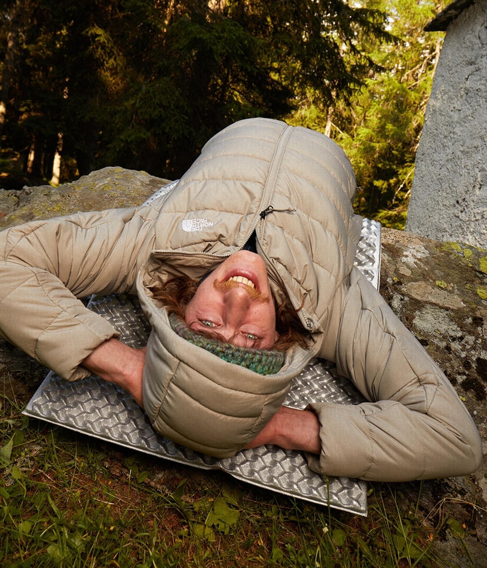 A person wearing a beige North Face puffer jacket smiles upside down while lying on a sleeping pad in a forest.