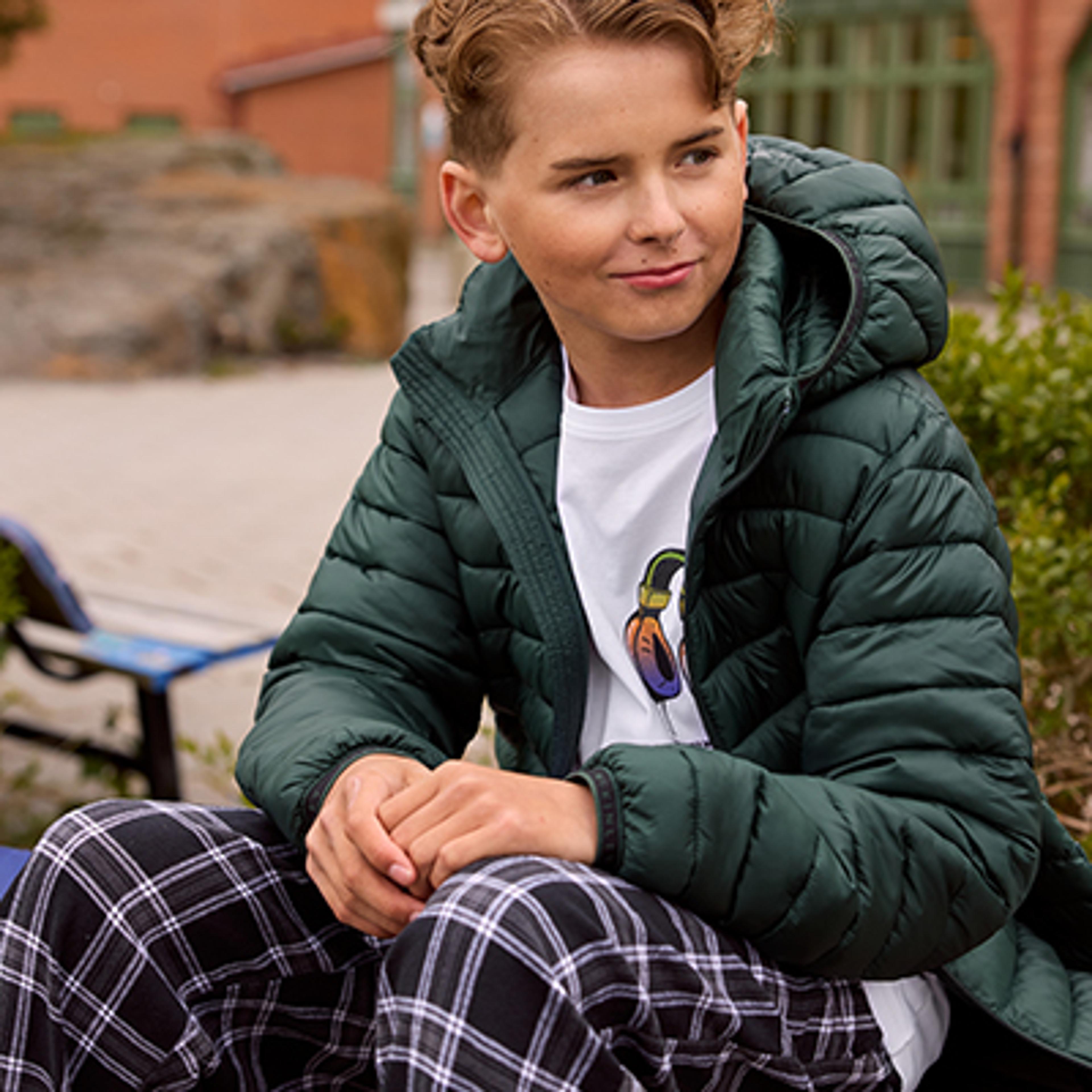 a young boy wearing a green jacket and plaid pants is sitting on a bench .