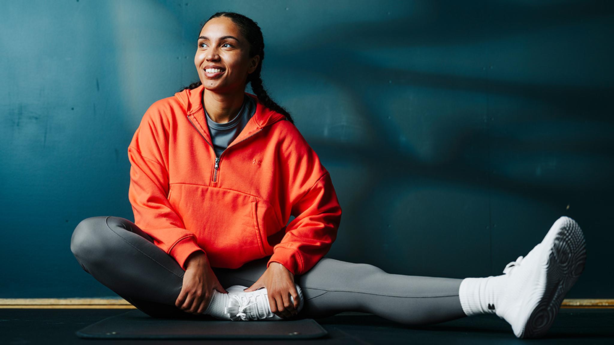 Smiling woman in an orange hoodie and grey leggings stretching on a mat.