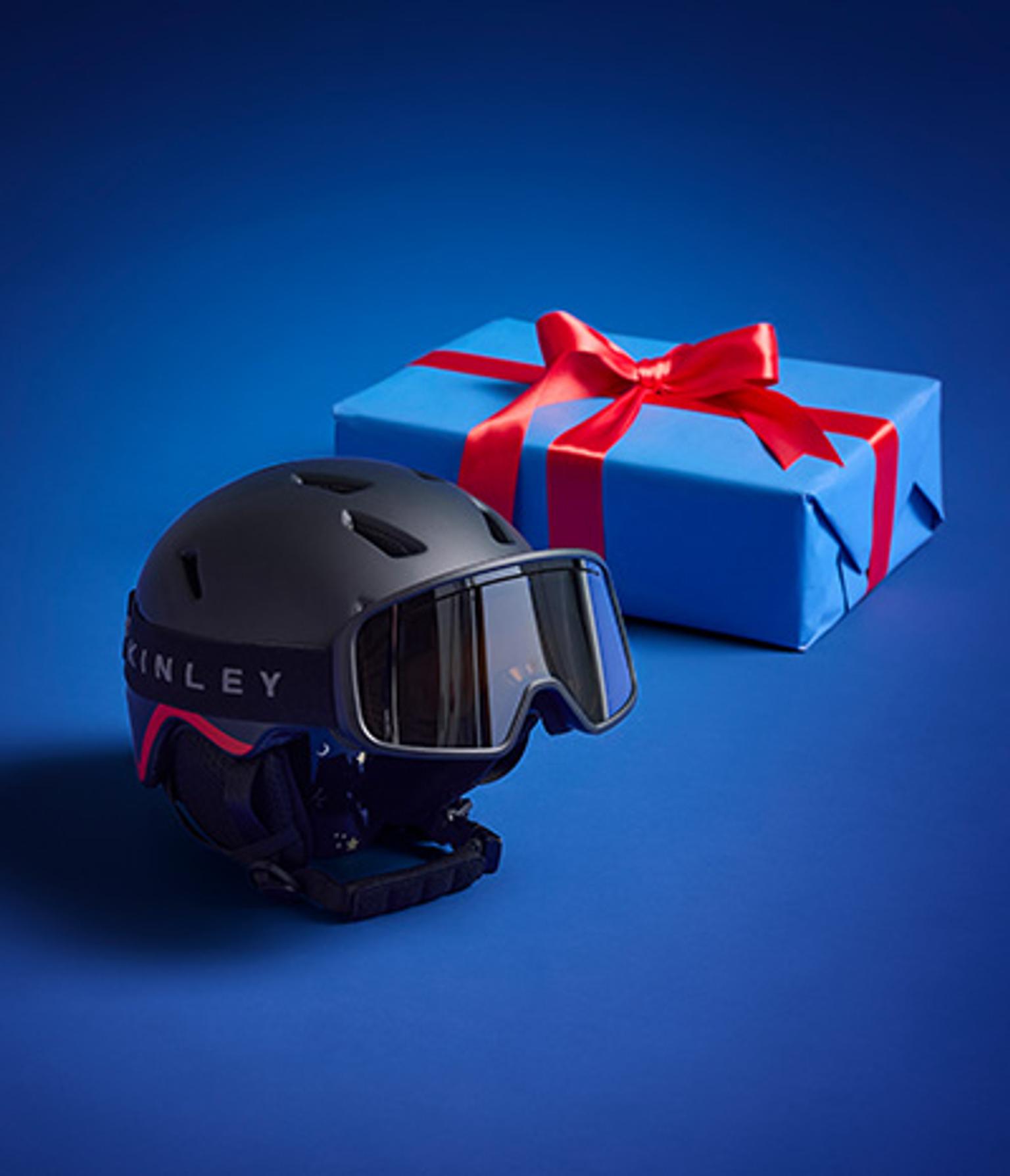 Dark grey ski helmet and goggles in front of a blue gift box with a red ribbon.