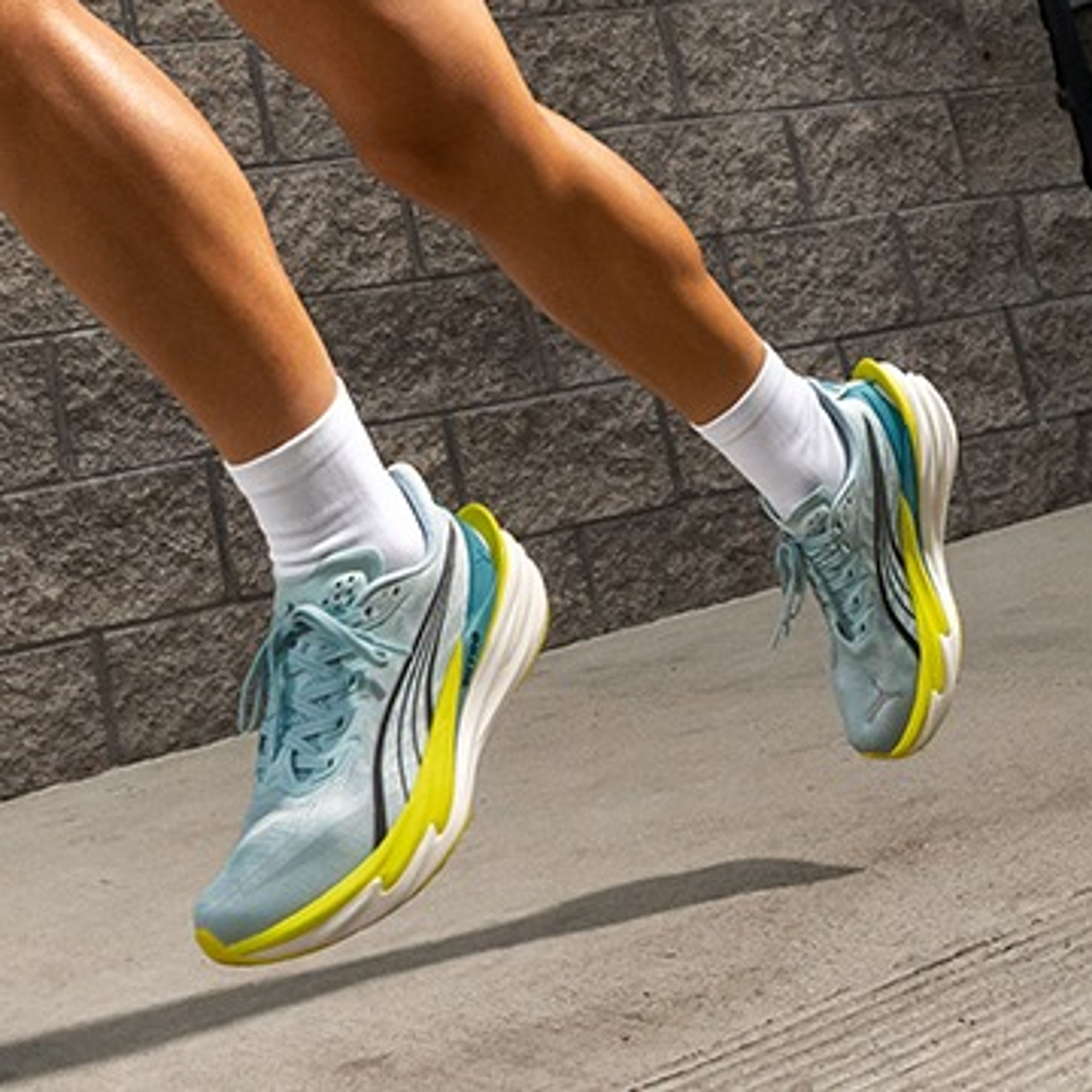 A woman in a light blue athletic outfit and blue-yellow running shoes runs energetically in an urban setting.