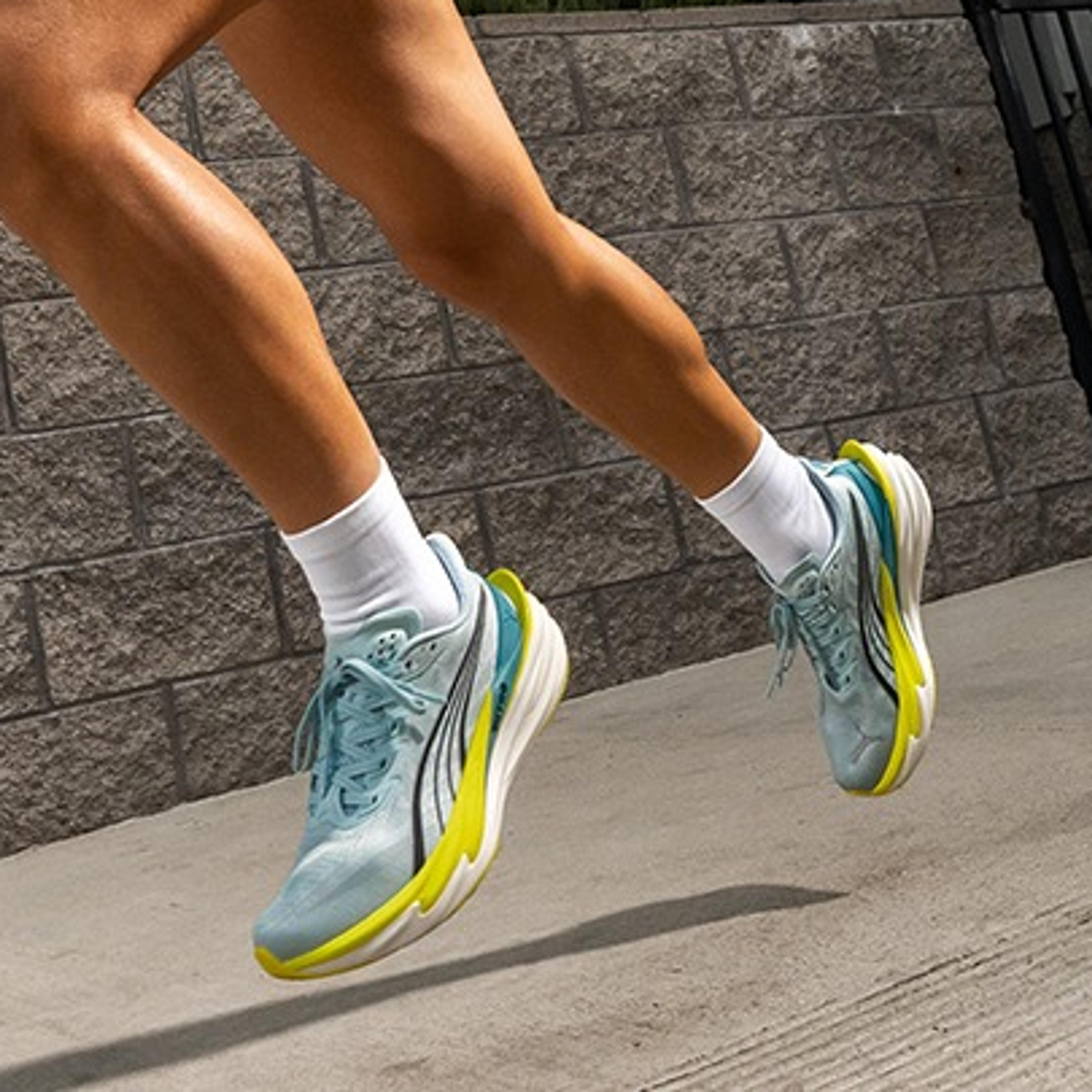 A woman runs outdoors in a light blue Puma athletic top, shorts, and light blue and yellow sneakers.