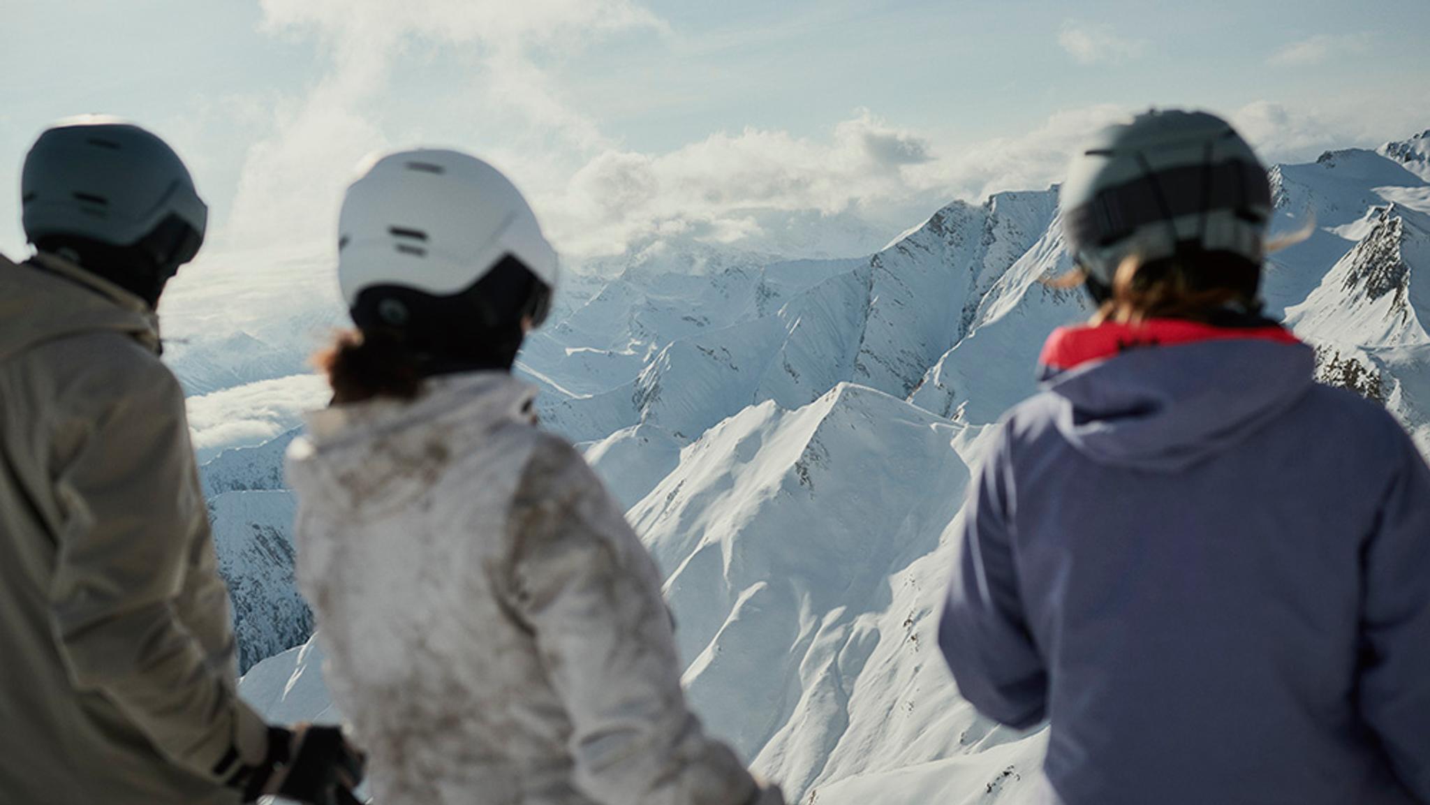 Three skiers, seen from behind, look out at a vast snow-covered mountain range.
