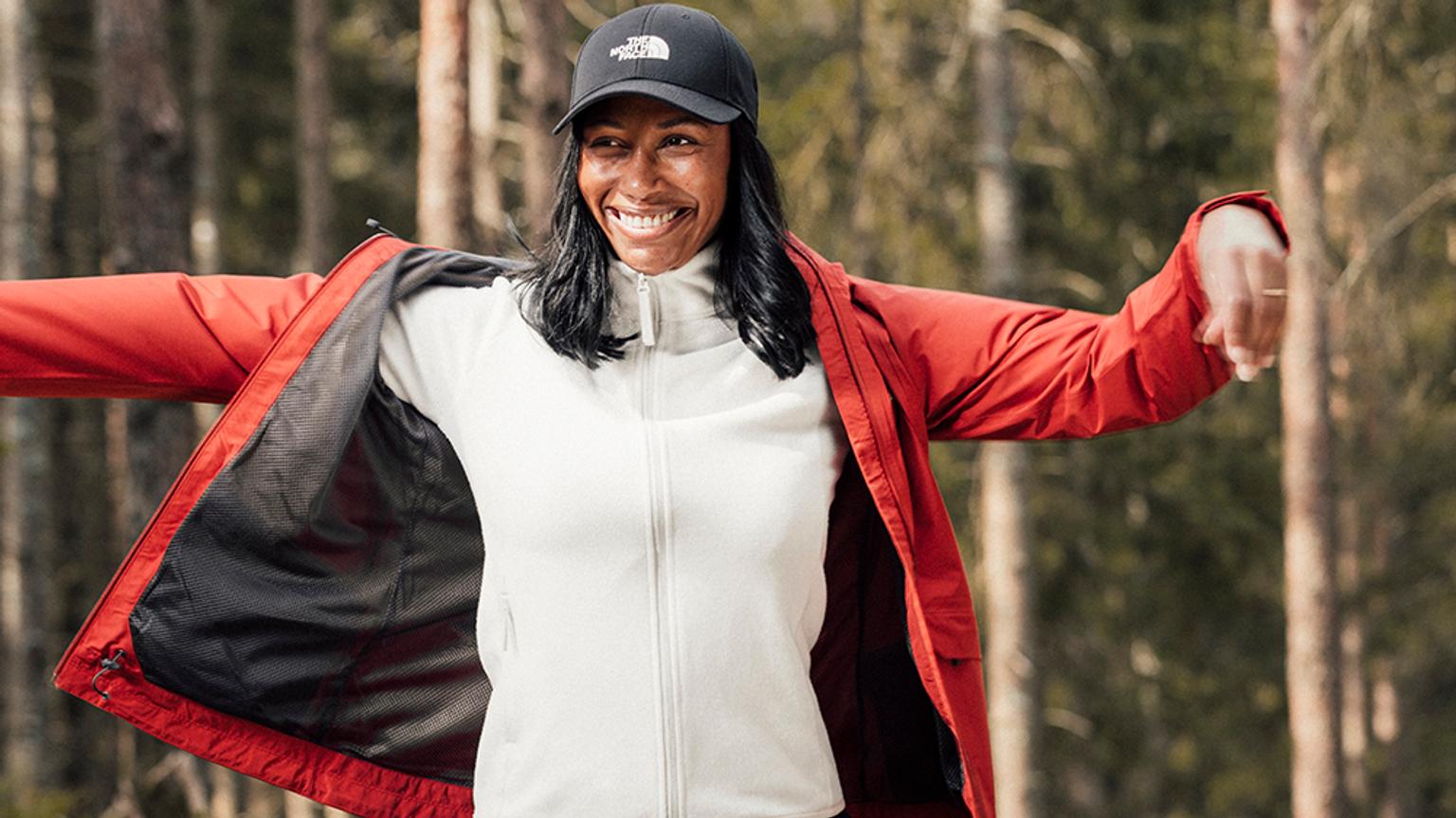 a woman wearing a red jacket and a black hat is standing in the woods with her arms outstretched .