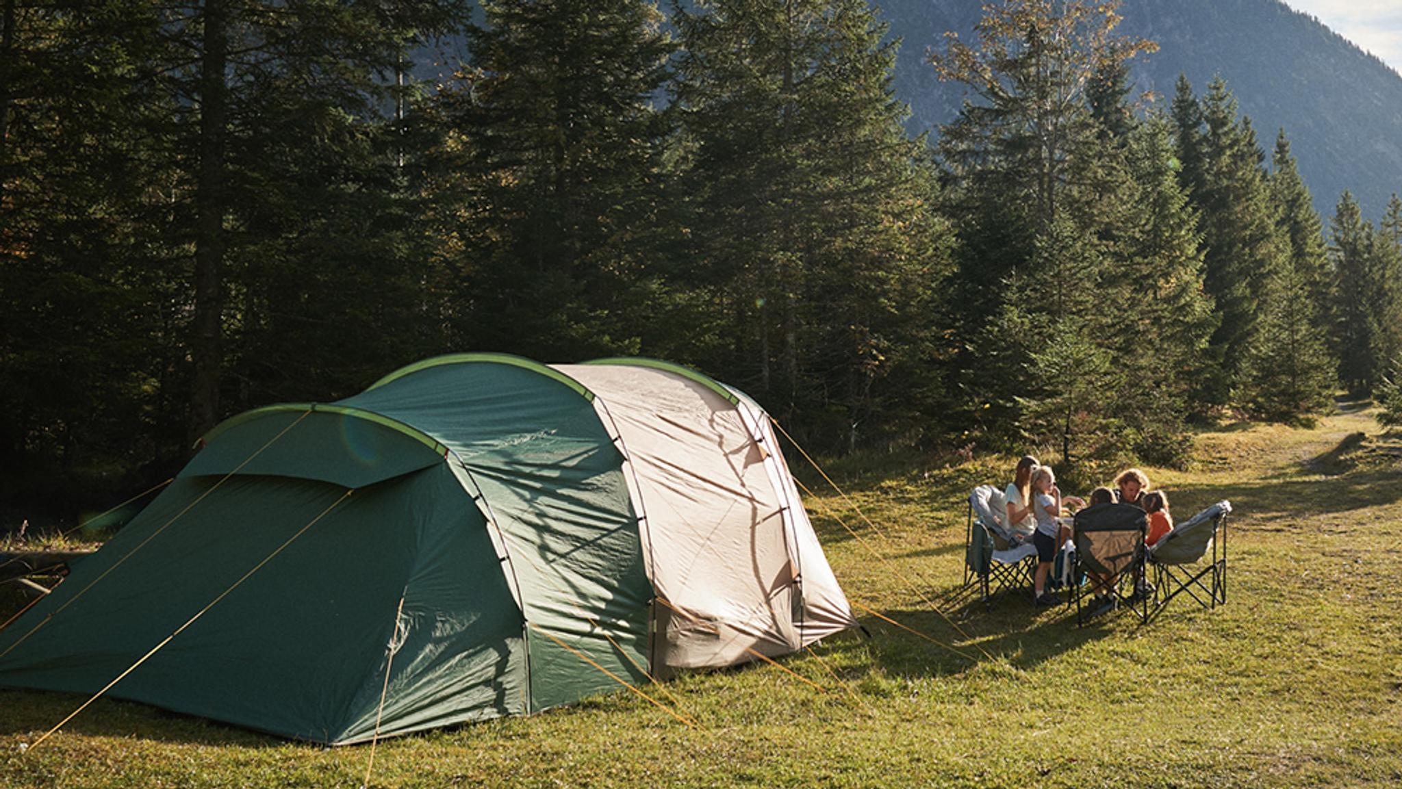 A family sits around a table outside a large tent in a sunny forest.