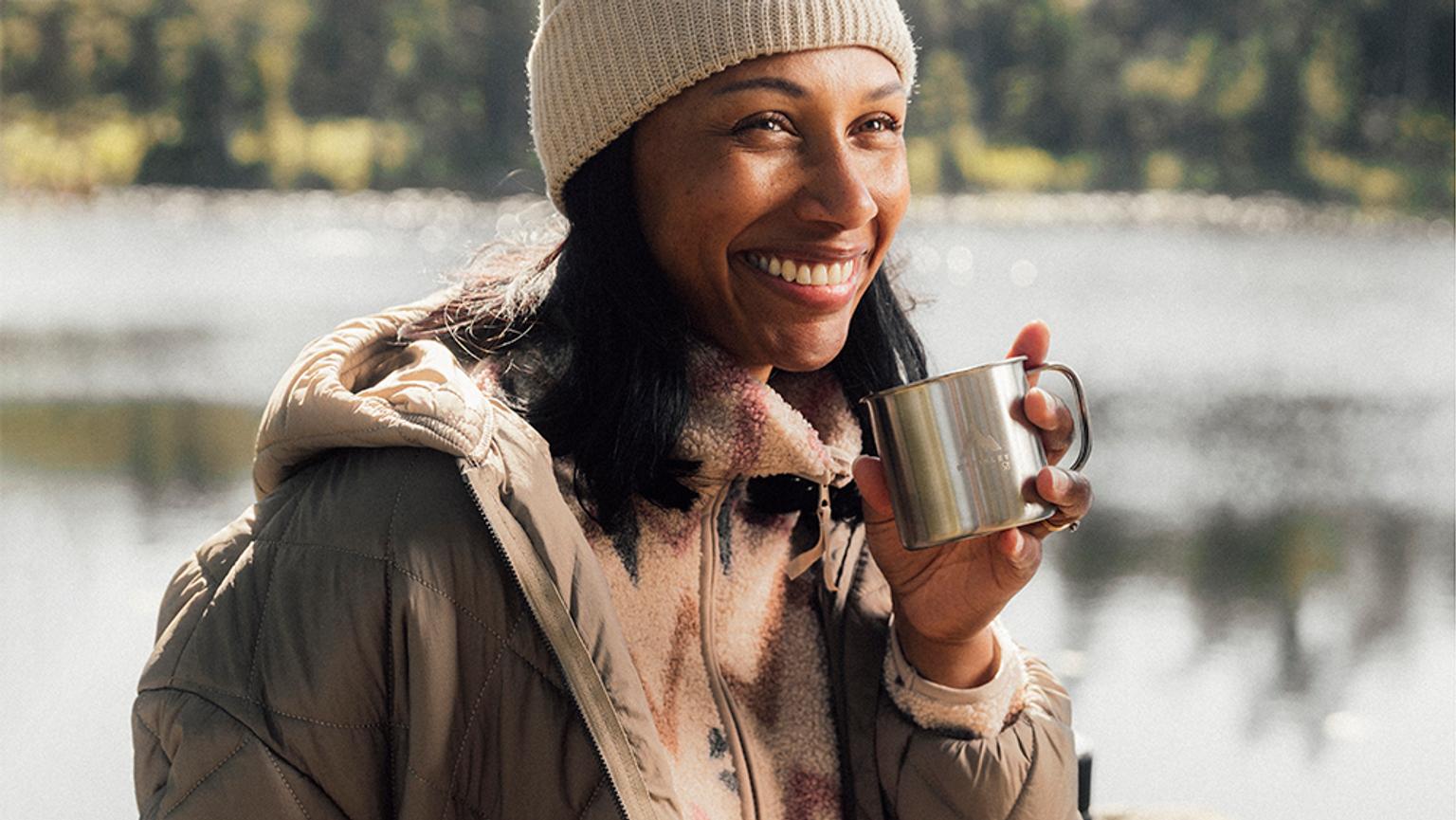 a woman is wearing a jacket and holding a cup of coffee in front of a lake .