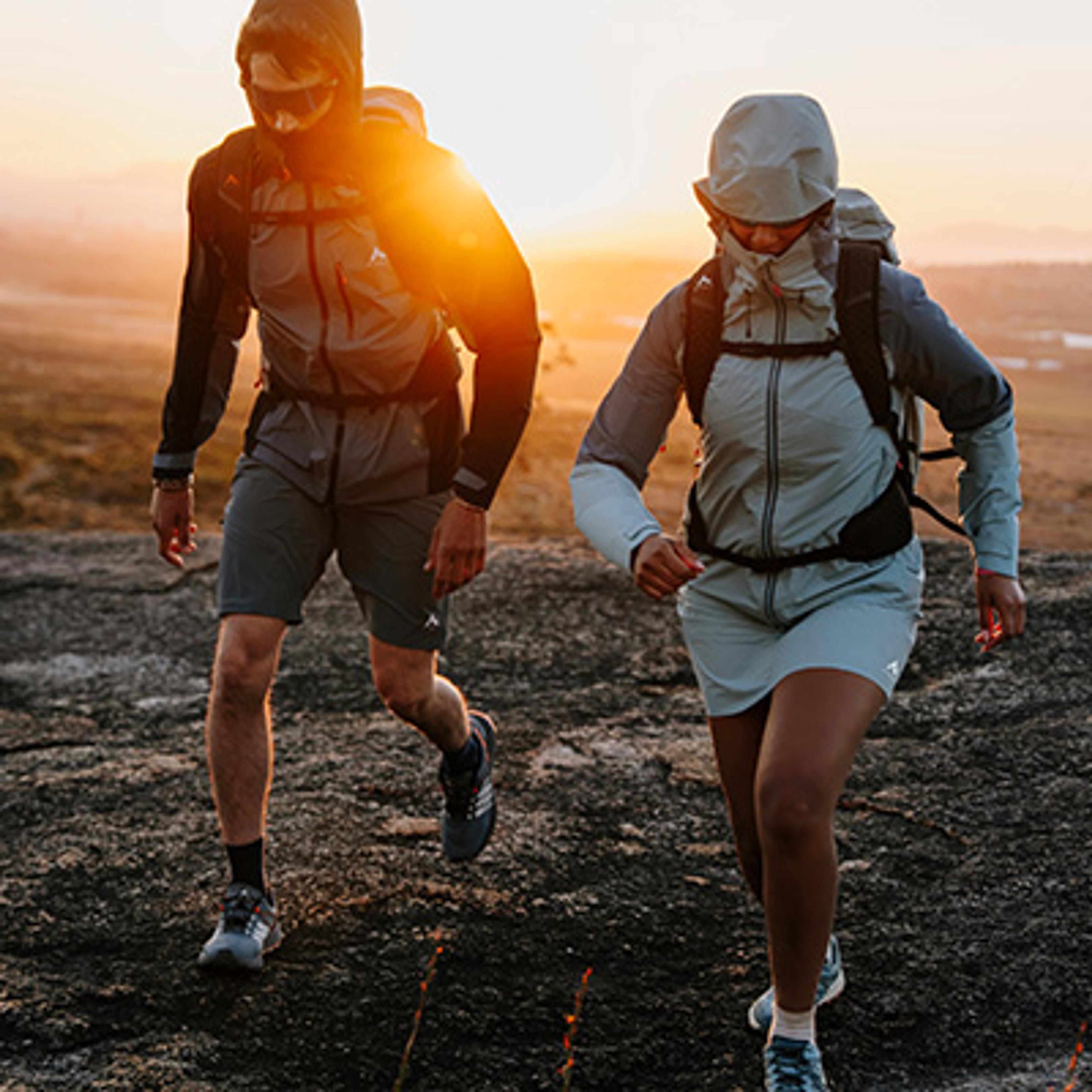 Two people in activewear and backpacks ascend a rocky trail at sunrise.
