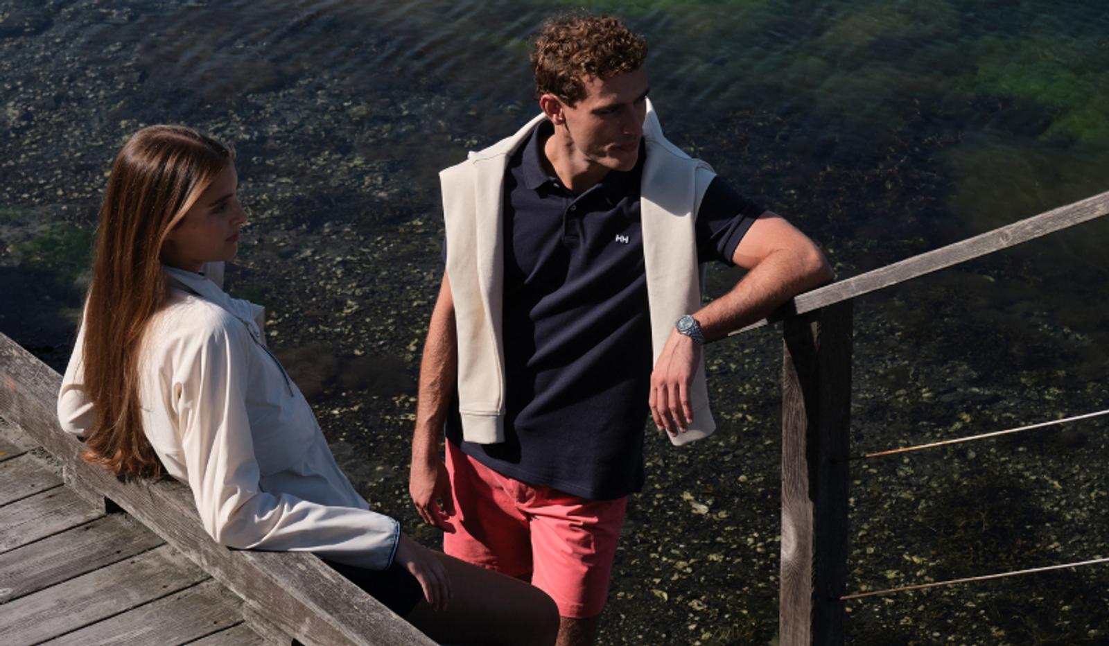 A young man stands while a young woman sits on a wooden dock by clear water.