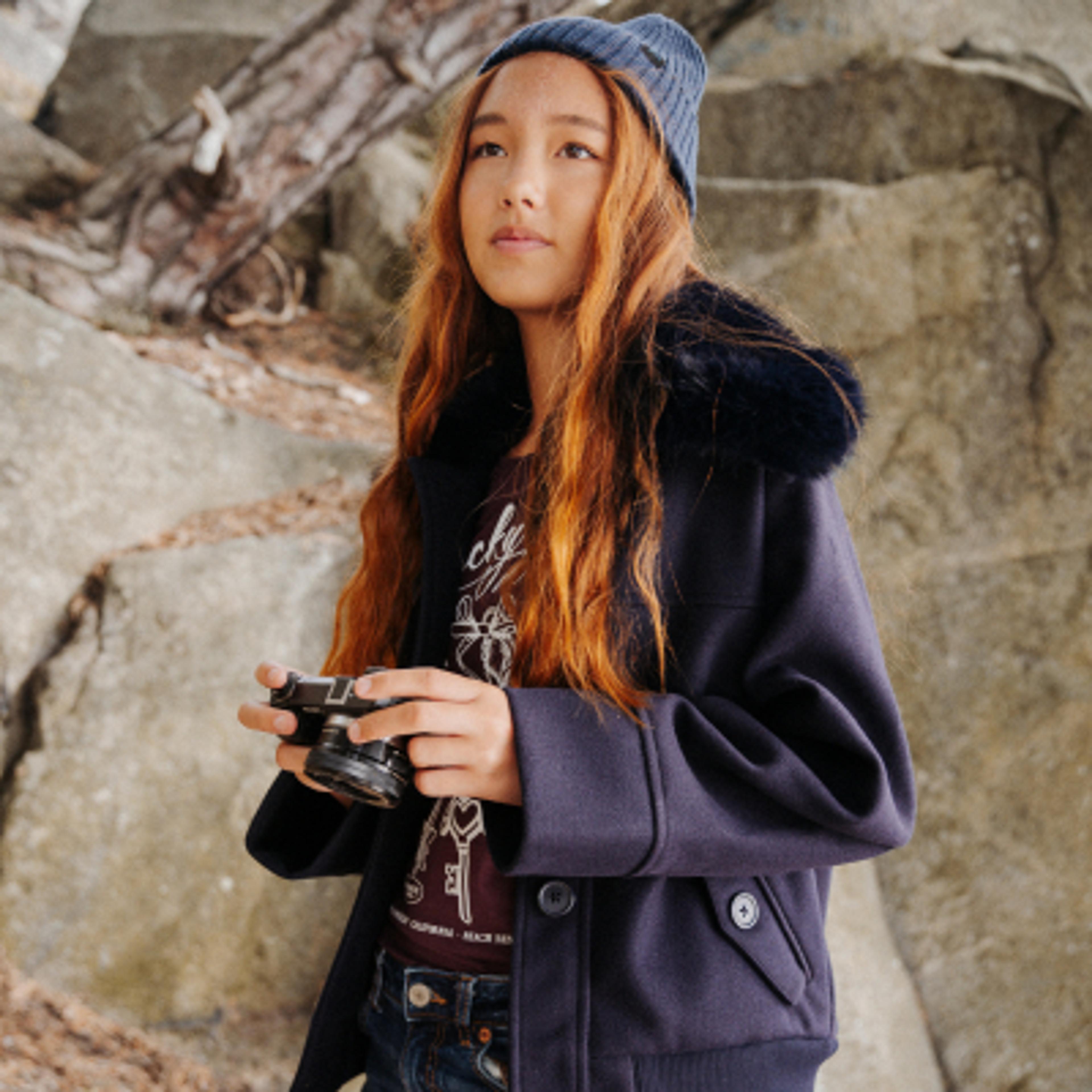 A young girl in a blue beanie and jacket holds a camera, looking thoughtfully outdoors.
