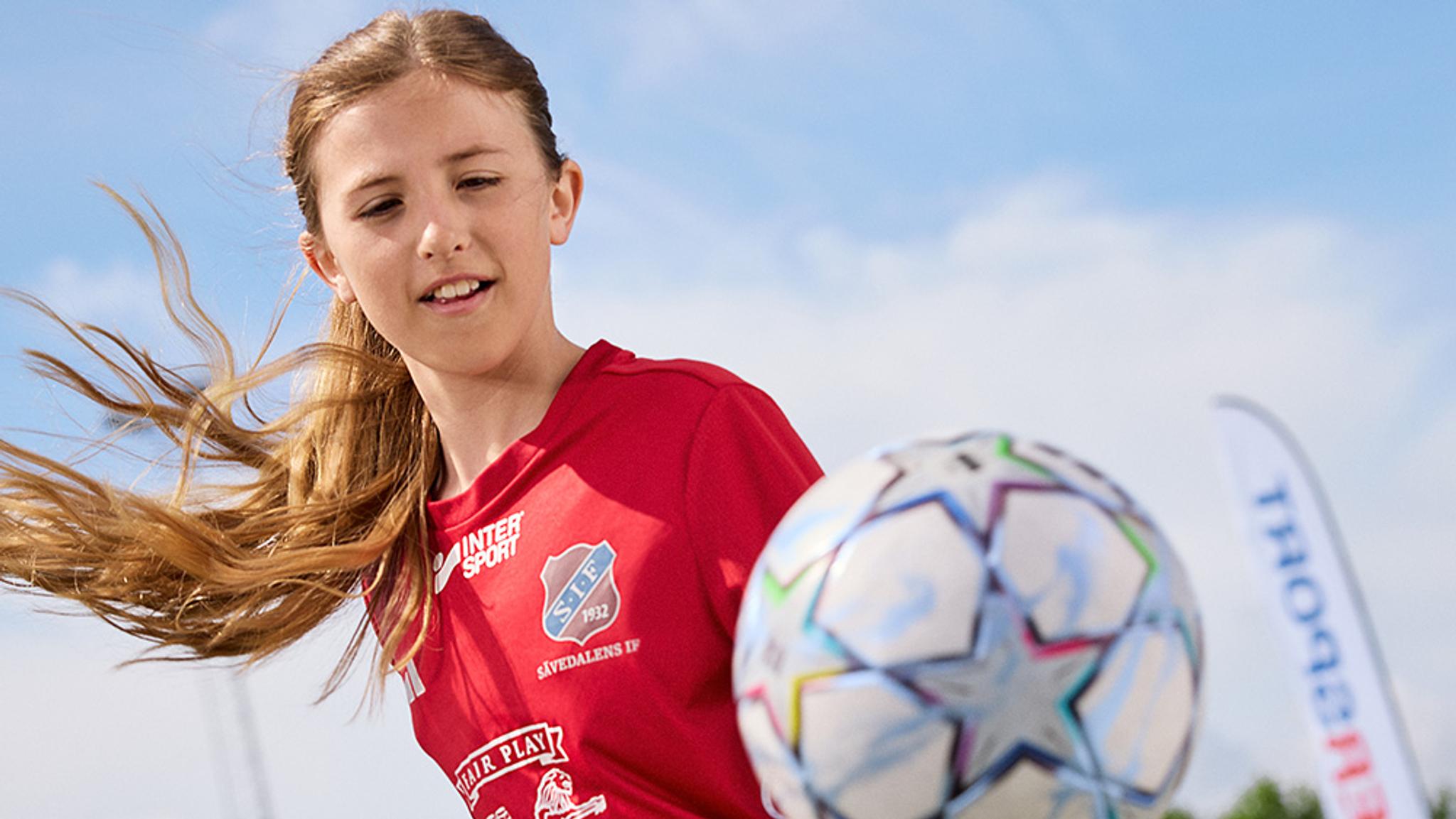 A young girl in a red jersey with wind-blown hair looks at a soccer ball under a blue sky.