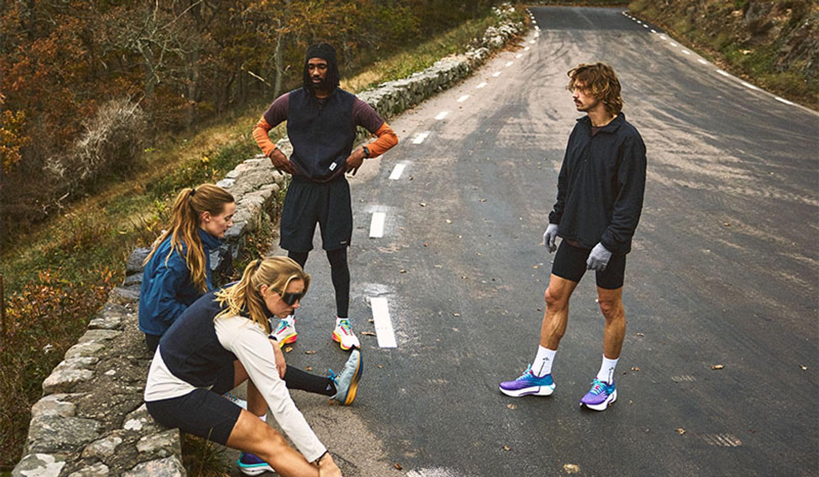 Four runners in athletic wear on a winding road, one stretching on a stone wall, another standing with hands on hips, and a third standing further down the road.