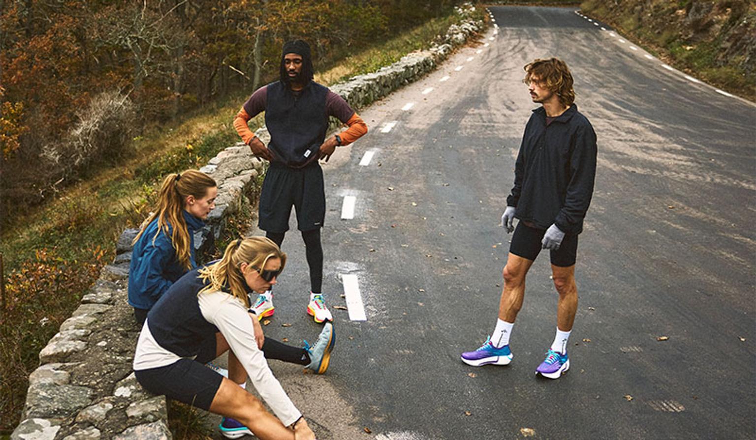 Four runners in athletic wear on a winding road, one stretching on a stone wall, another standing with hands on hips, and a third standing further down the road.