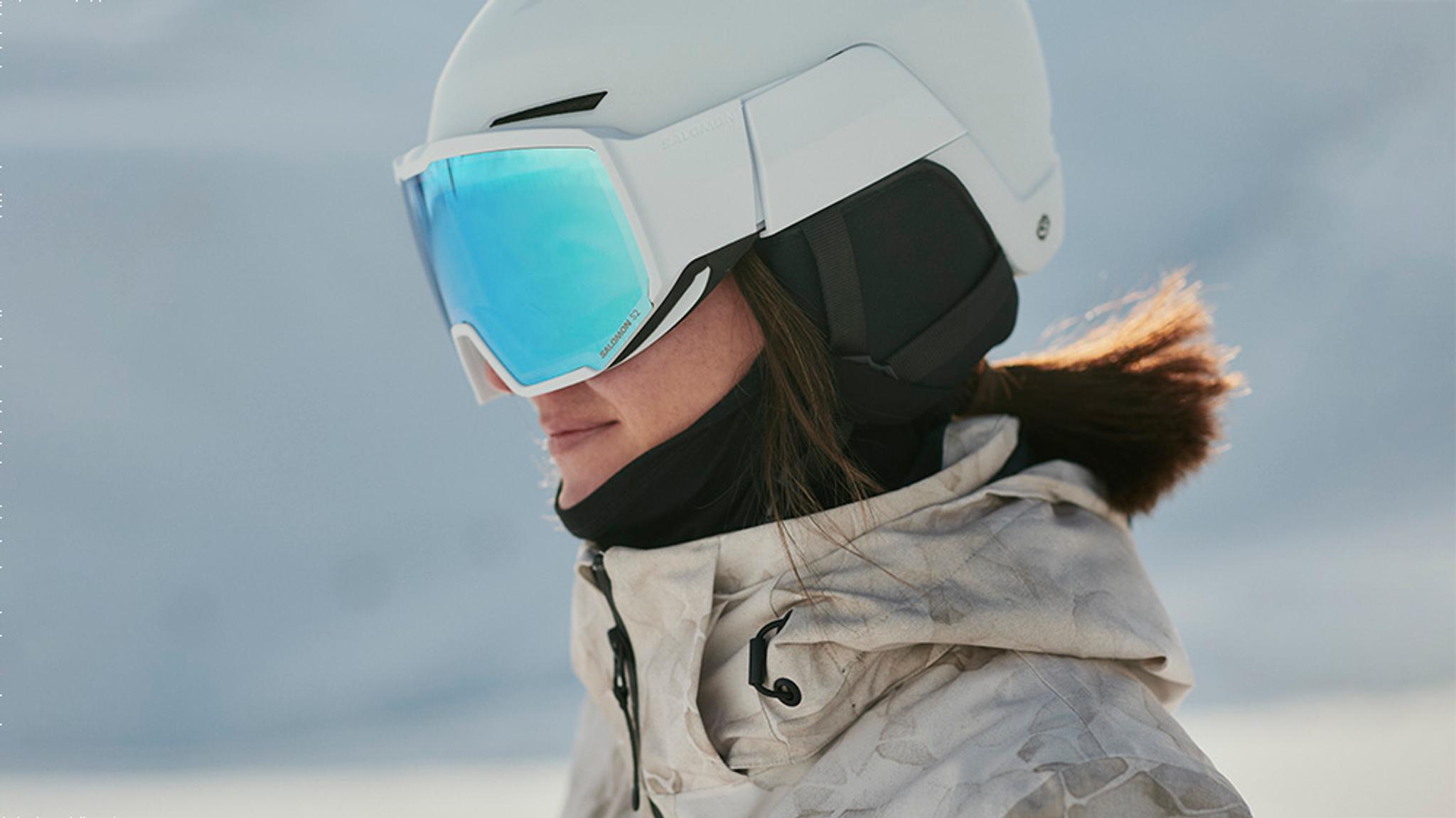 Side profile of a person wearing a white ski helmet, blue mirrored goggles, and a camouflage winter jacket.