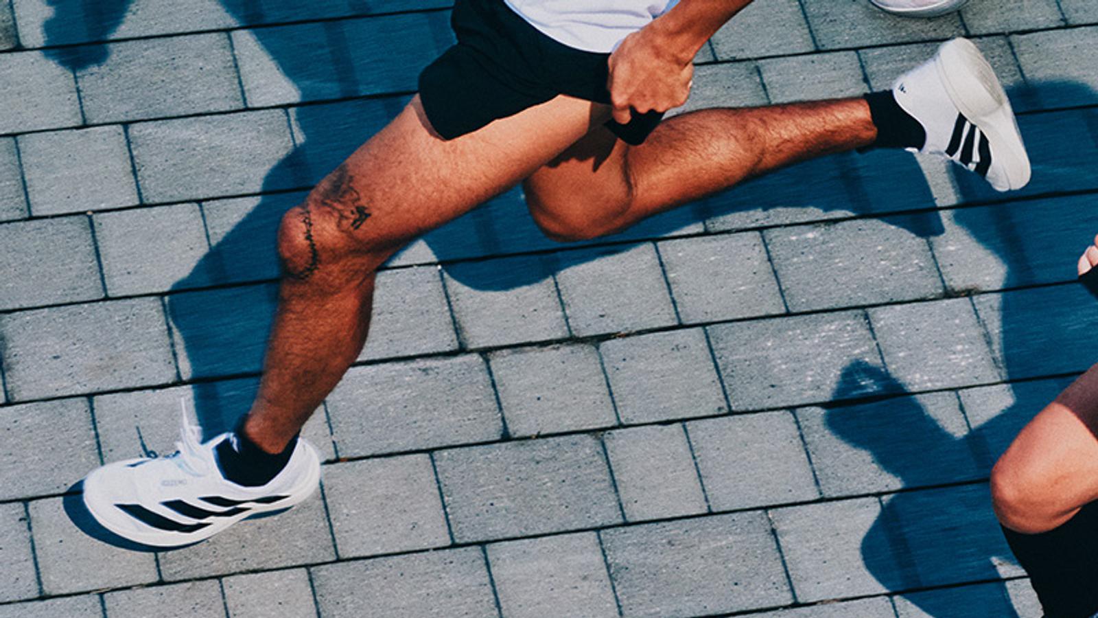 A person's legs, wearing white running shoes and black shorts, running on grey paved bricks. A tattoo is visible on one knee.