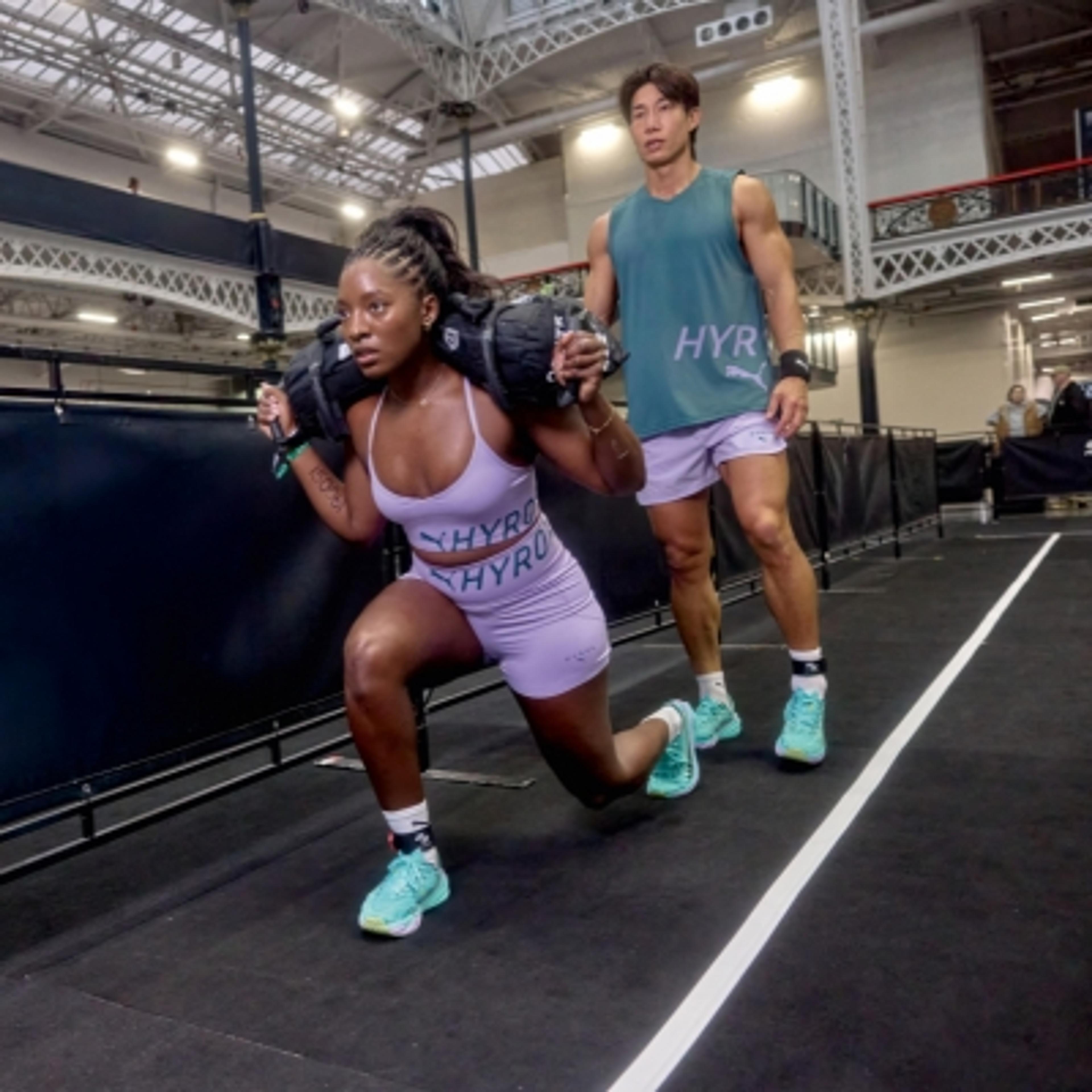 A woman in a purple HYROX outfit performs a sandbag lunge in a gym, while a man in a teal HYROX top watches.