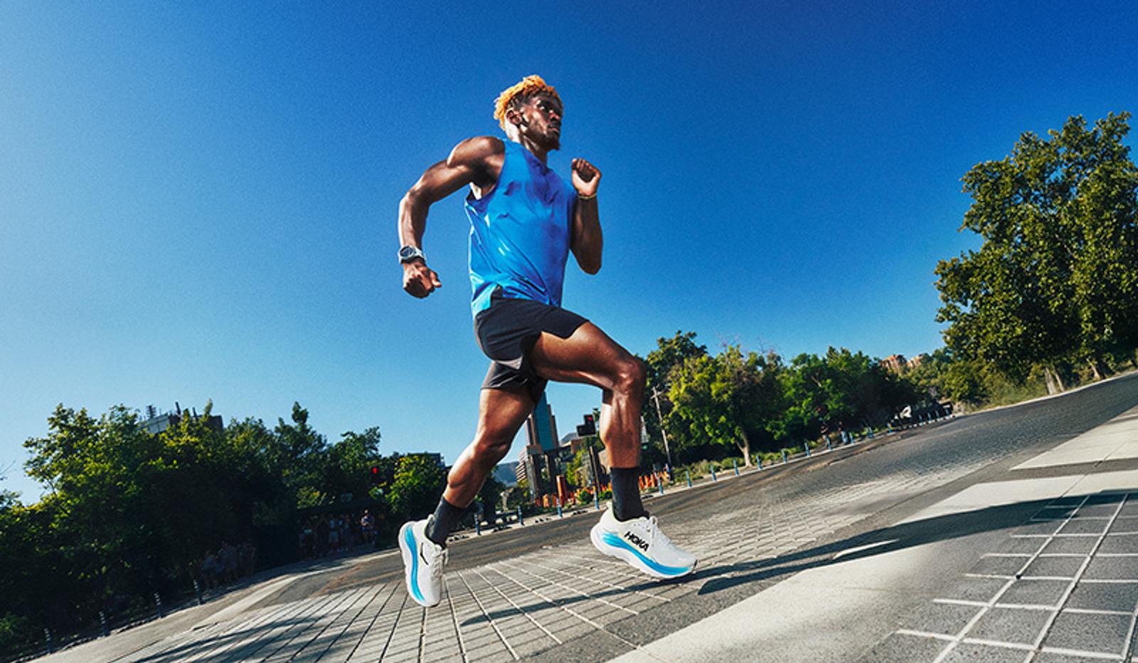 A male runner in a blue tank top and black shorts sprints outdoors on a sunny day.