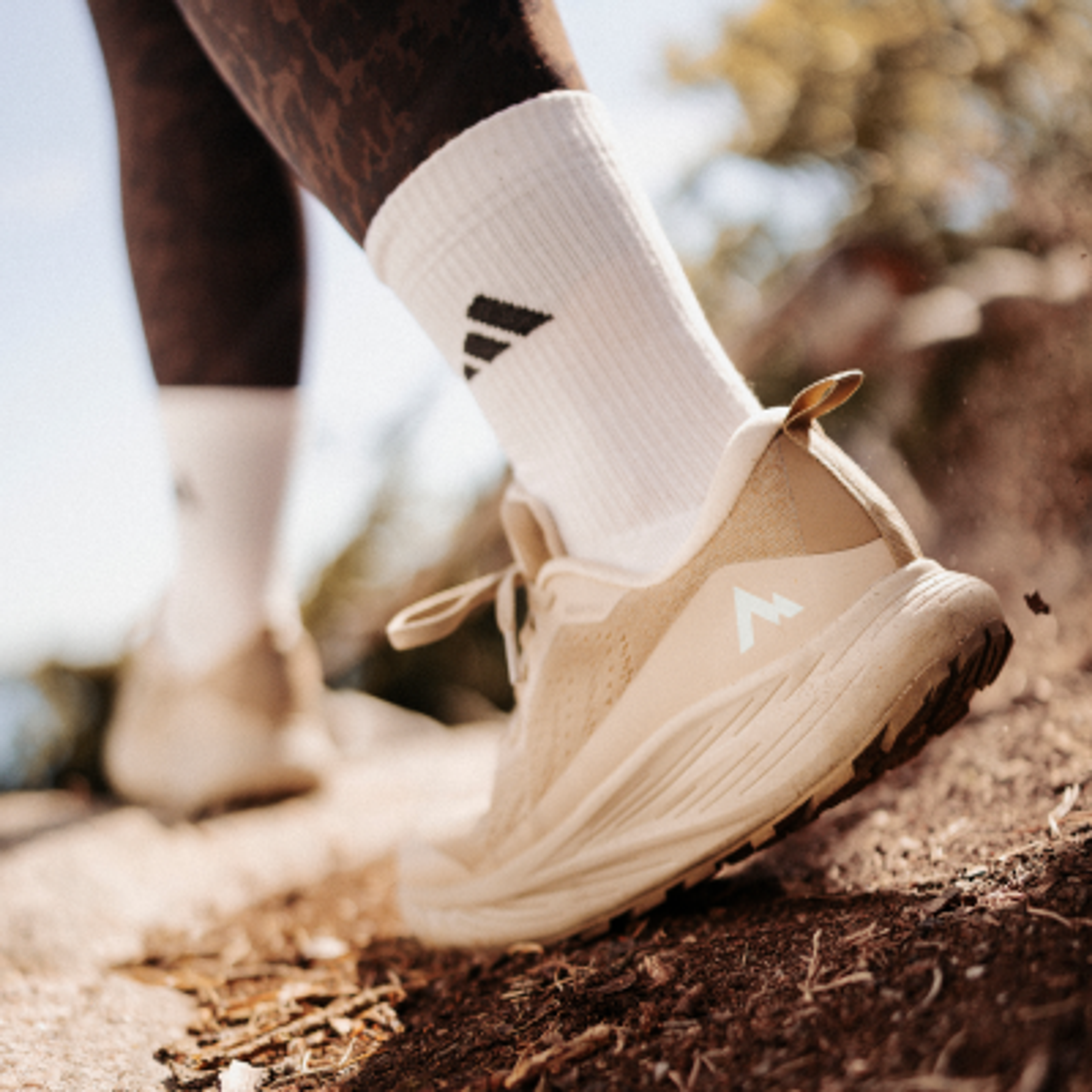 A foot in a beige trail shoe and white Adidas sock steps on a dirt path.