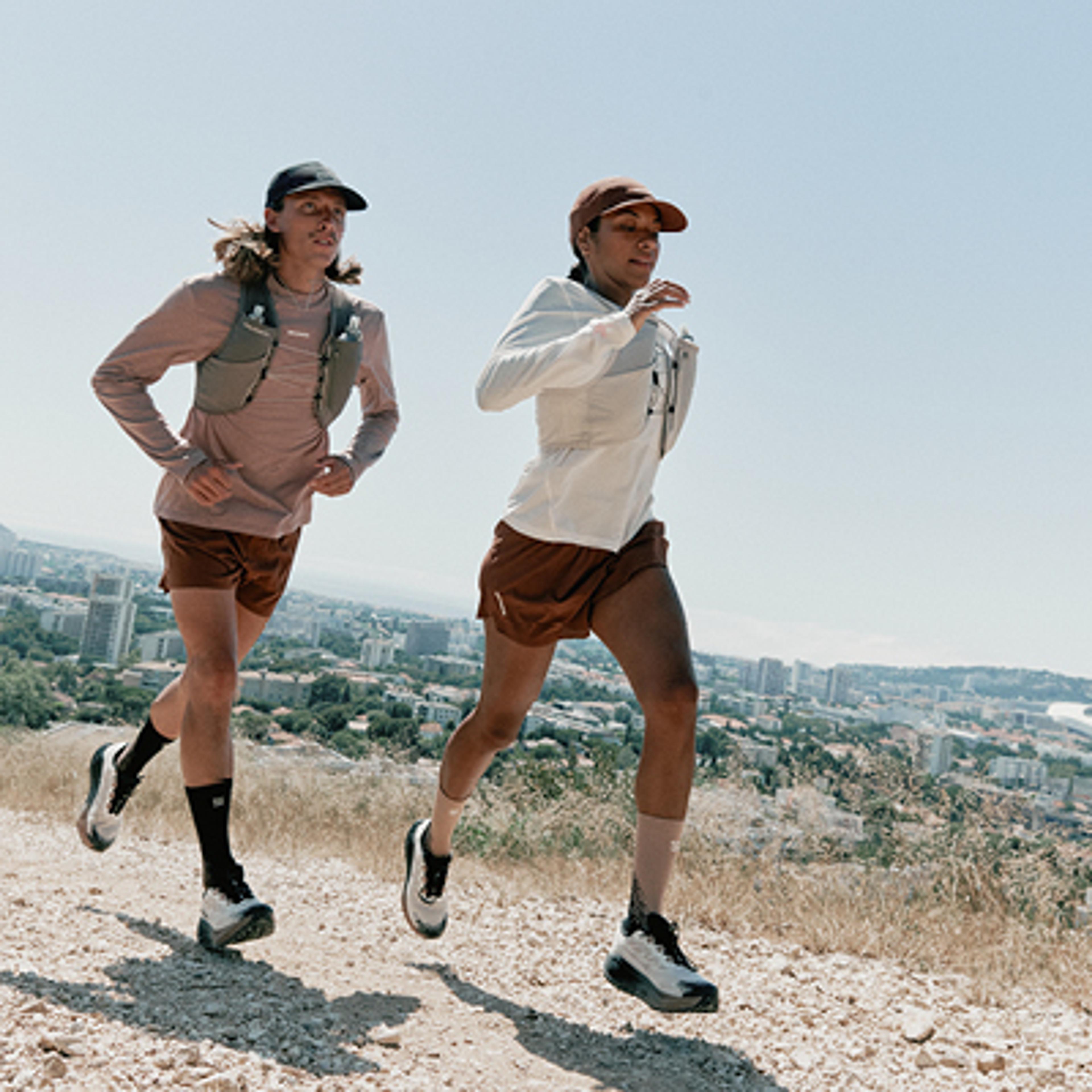 A man and a woman trail running on a dirt path with a city in the background.