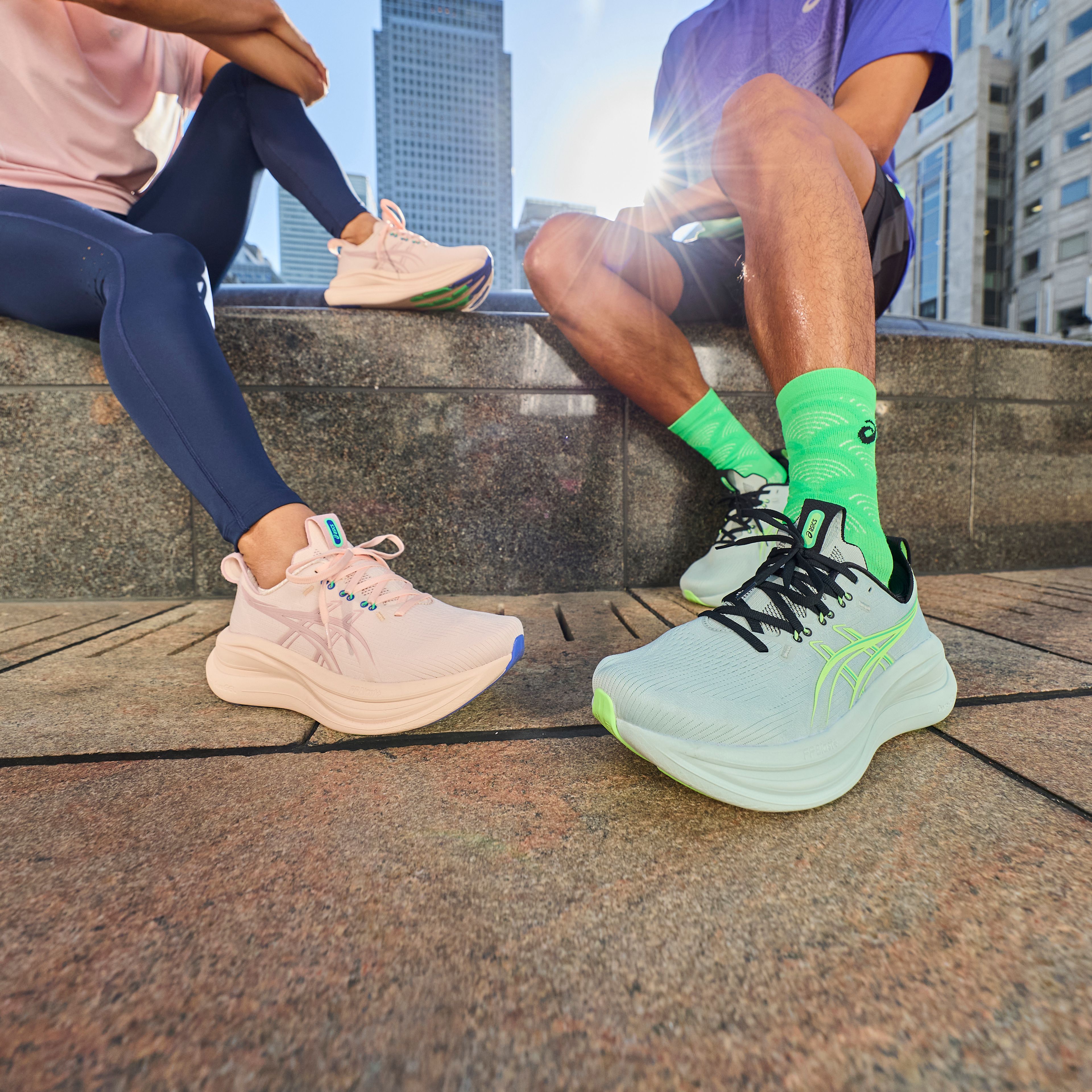 Two pairs of legs sitting in a city, wearing pink and light blue sneakers.