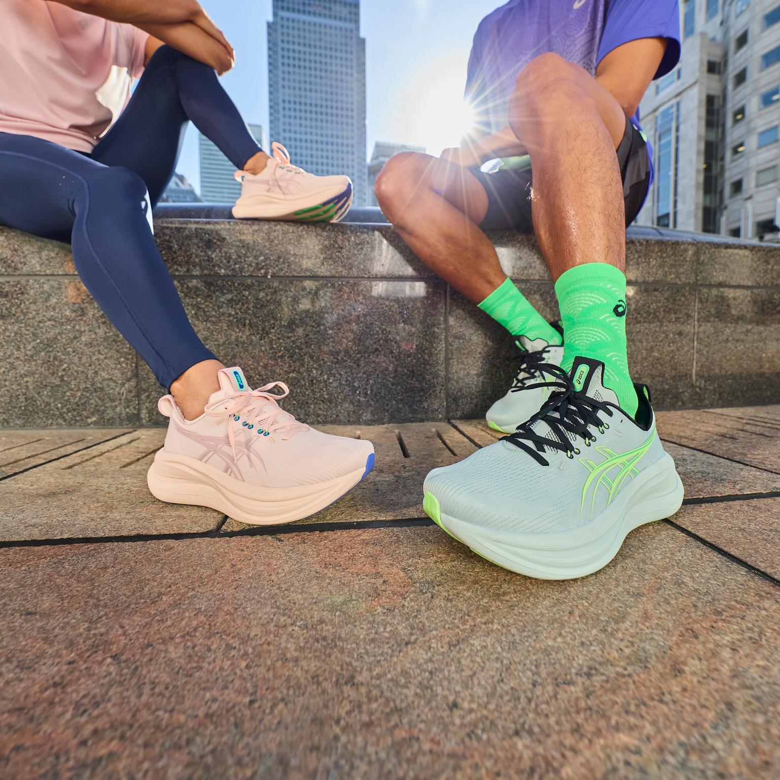 Two pairs of legs sitting in a city, wearing pink and light blue sneakers.