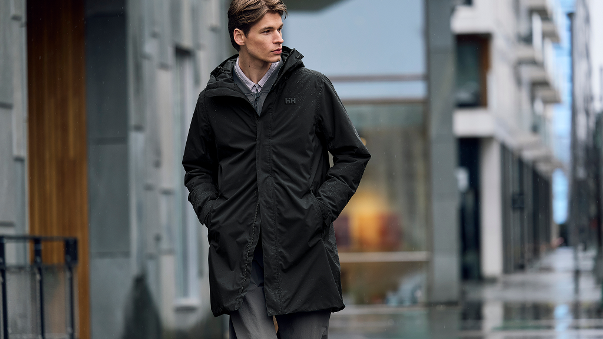 A man in a black hooded parka stands on a wet city street.