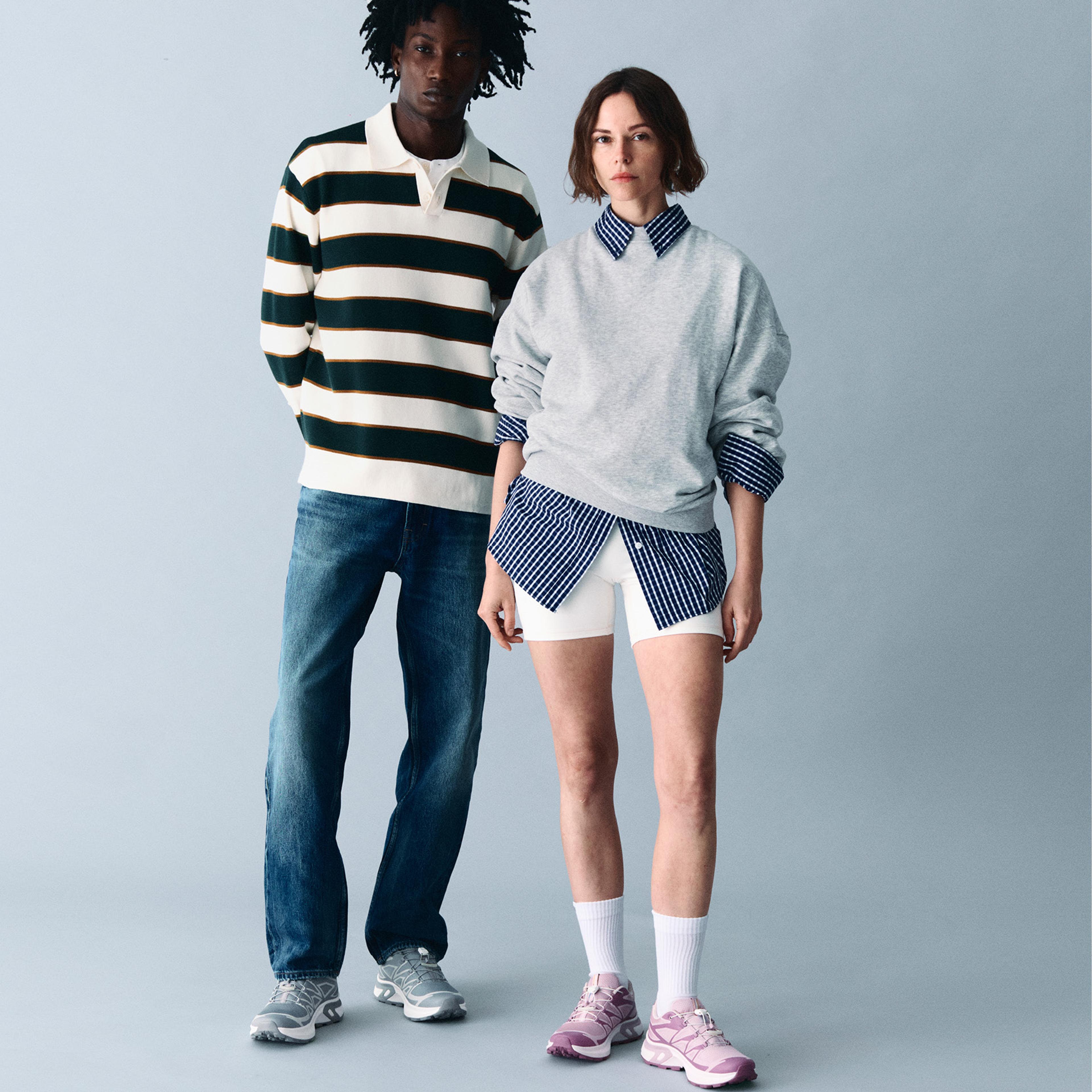 A man in a striped rugby shirt and jeans stands next to a woman in a grey sweatshirt over a pinstriped shirt and white shorts, both wearing sneakers.