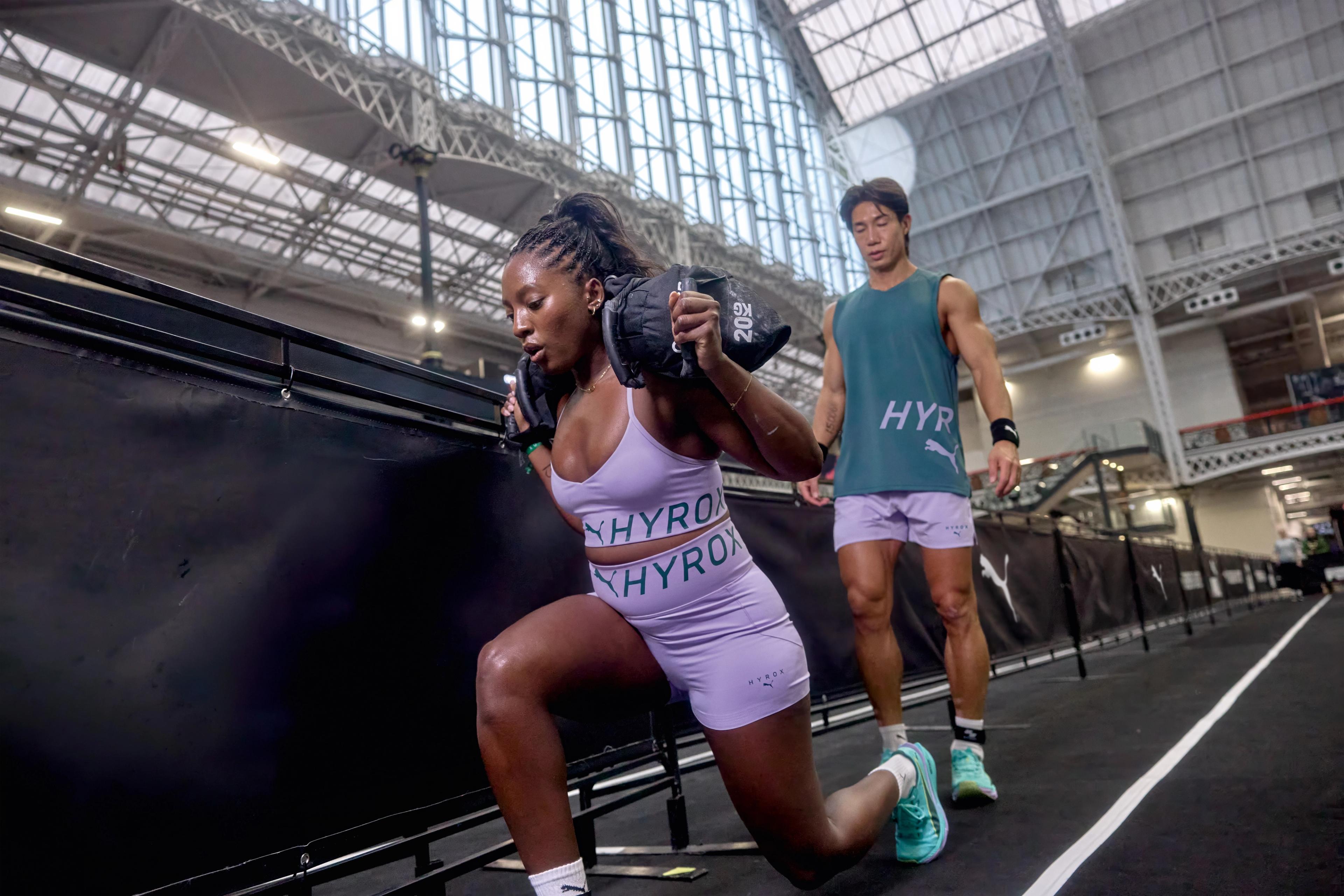 A woman performs a weighted lunge while a man watches in a large HYROX training facility.