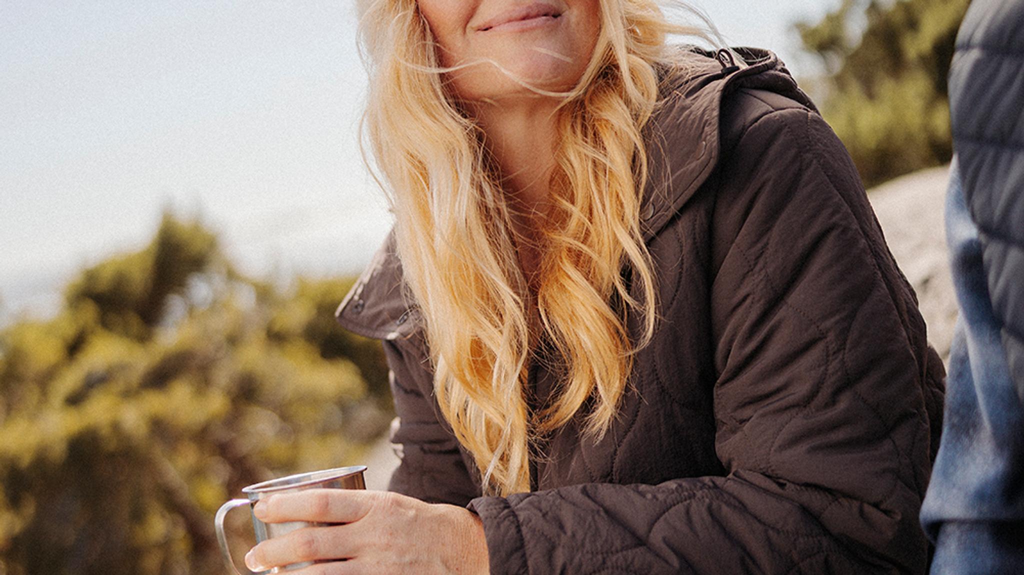 A woman with blonde hair, wearing a quilted jacket, holds a metal mug outdoors.