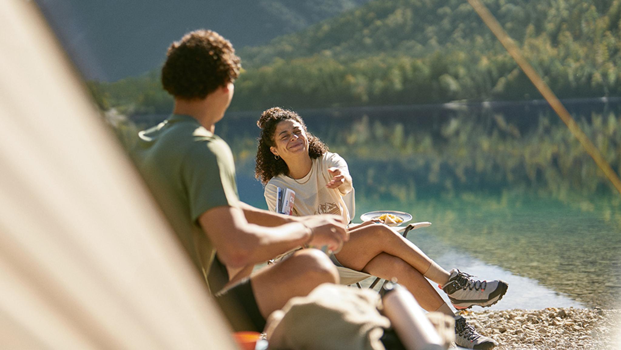 A woman smiles and gestures towards a man while relaxing by a scenic lake with mountains.