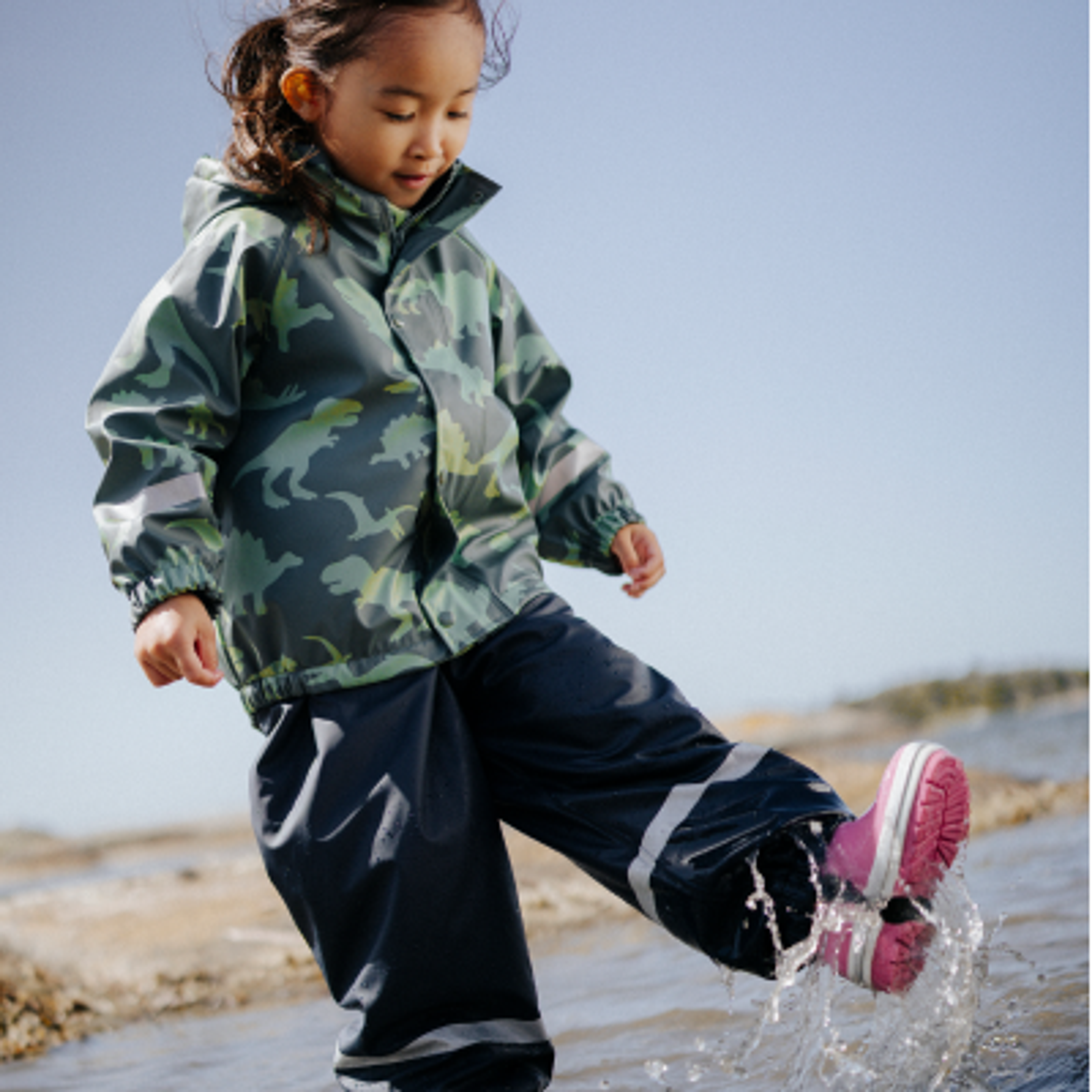 A child in a dinosaur-patterned rain jacket and pink boots splashes water.