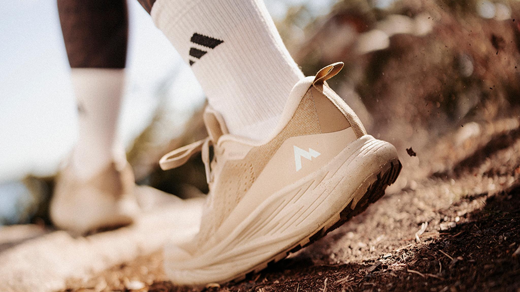 Close-up of a beige trail shoe with a white Adidas sock stepping on a dirt path, kicking up dust.