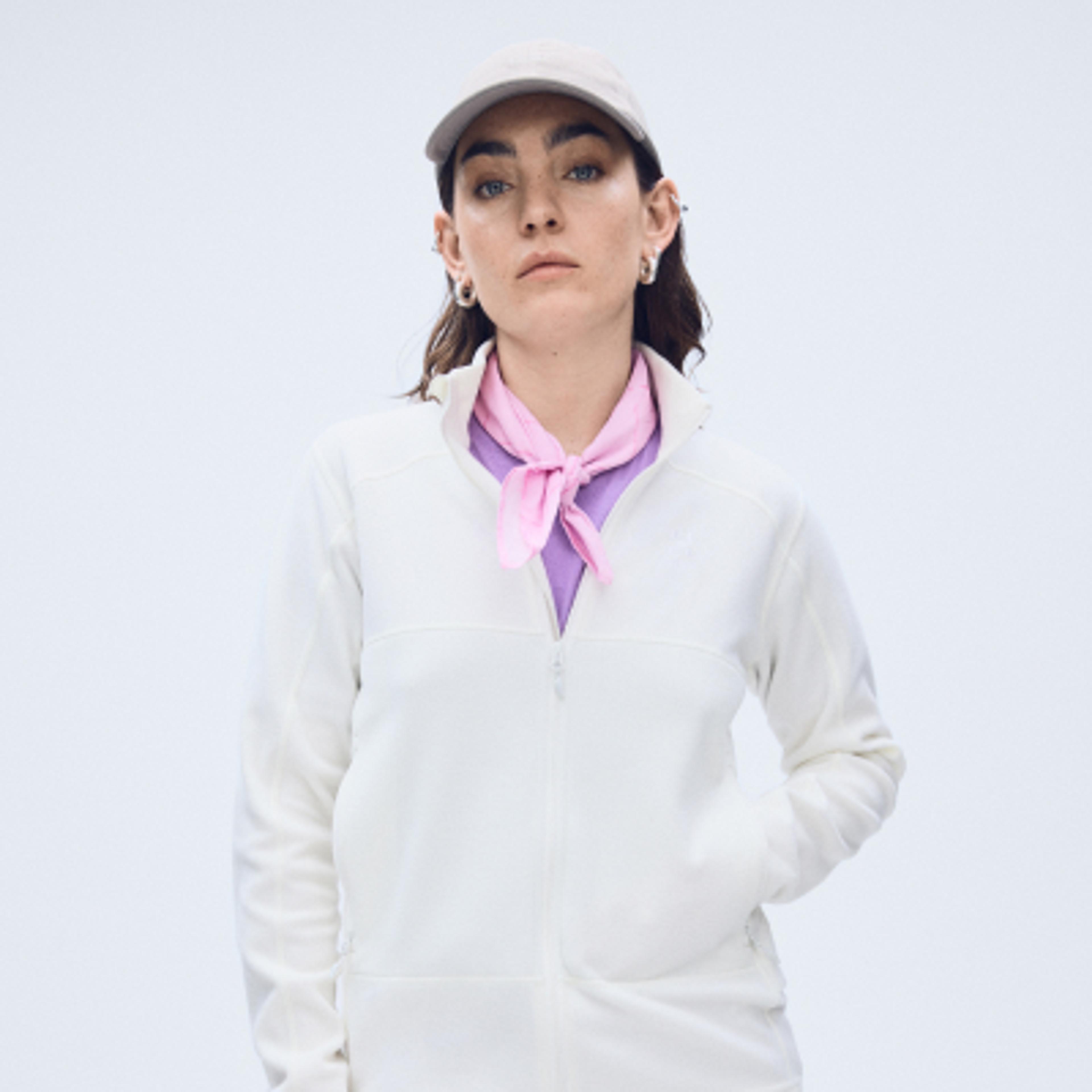 A woman in a light cap, white fleece jacket, purple top, and pink bandana, looking forward with her hand in her pocket.
