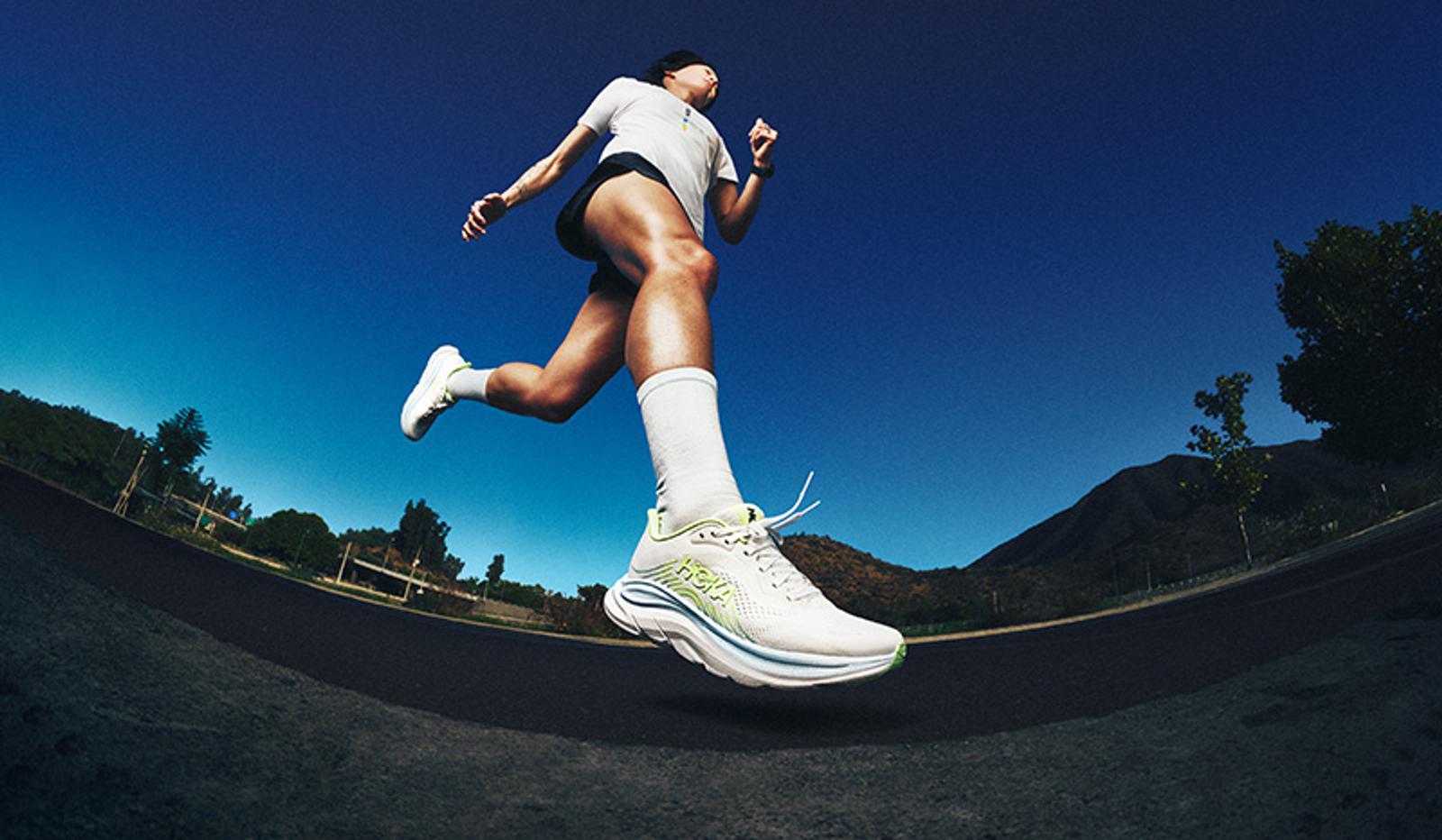 Low-angle shot of a person running mid-stride, wearing white and light green running shoes, against a blue sky.
