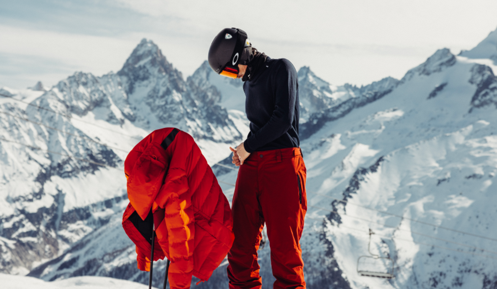 A skier in red pants and a helmet stands next to a red jacket on ski poles, with snowy mountains in the background.