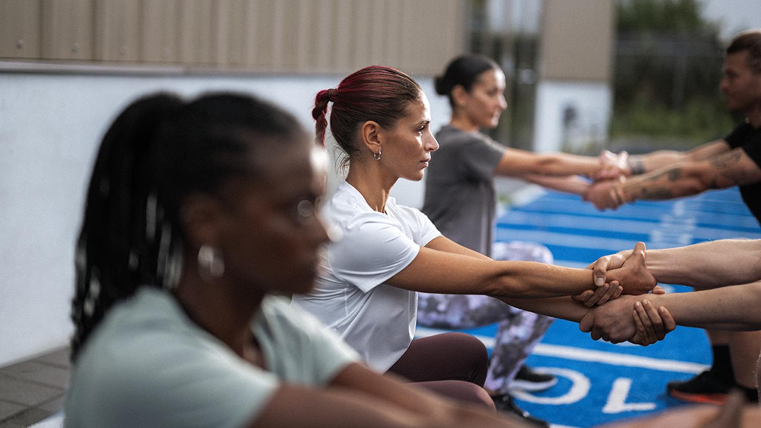 A diverse group of people performs assisted squats, holding each other's forearms, on an outdoor blue track.